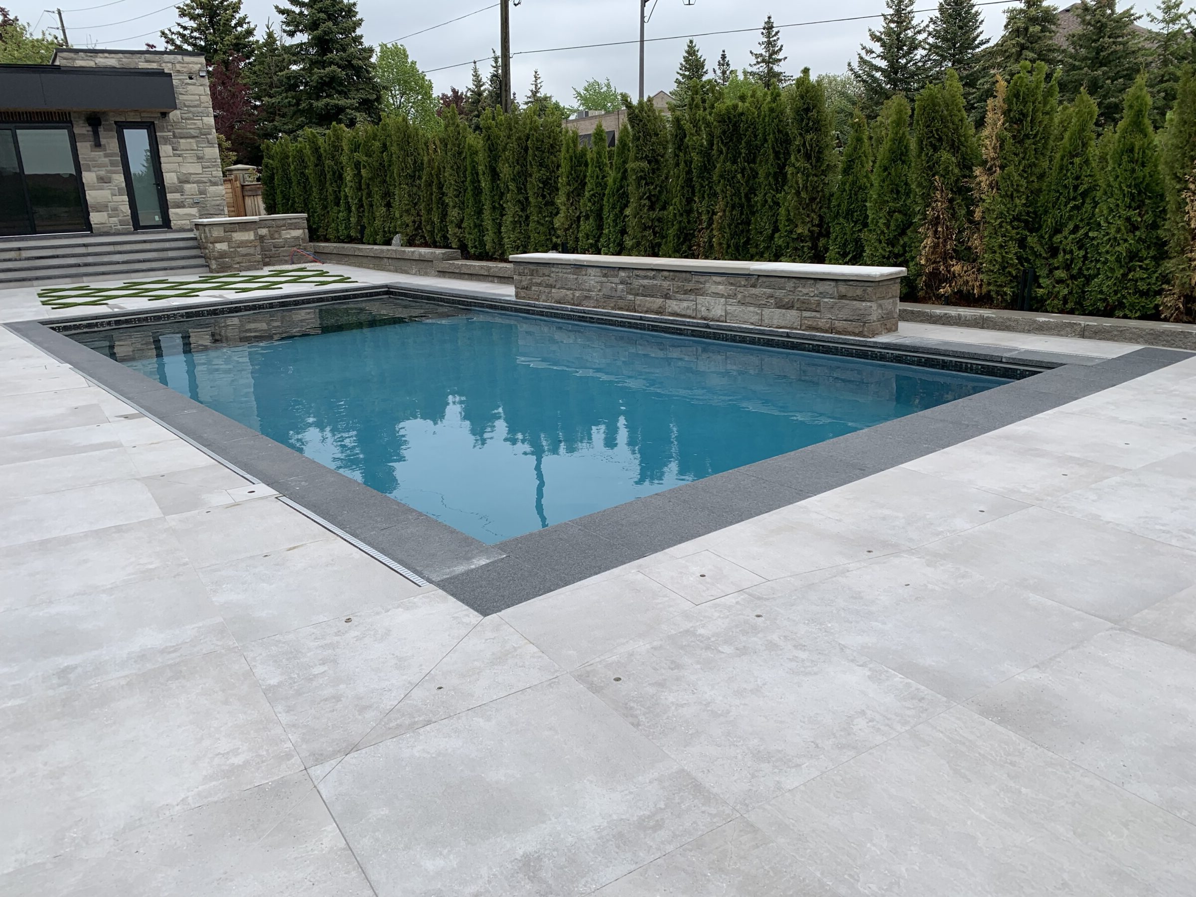 Modern outdoor pool with stone-bordered landscaping, surrounded by tall evergreen trees. Adjacent to a contemporary stone building under a cloudy sky.