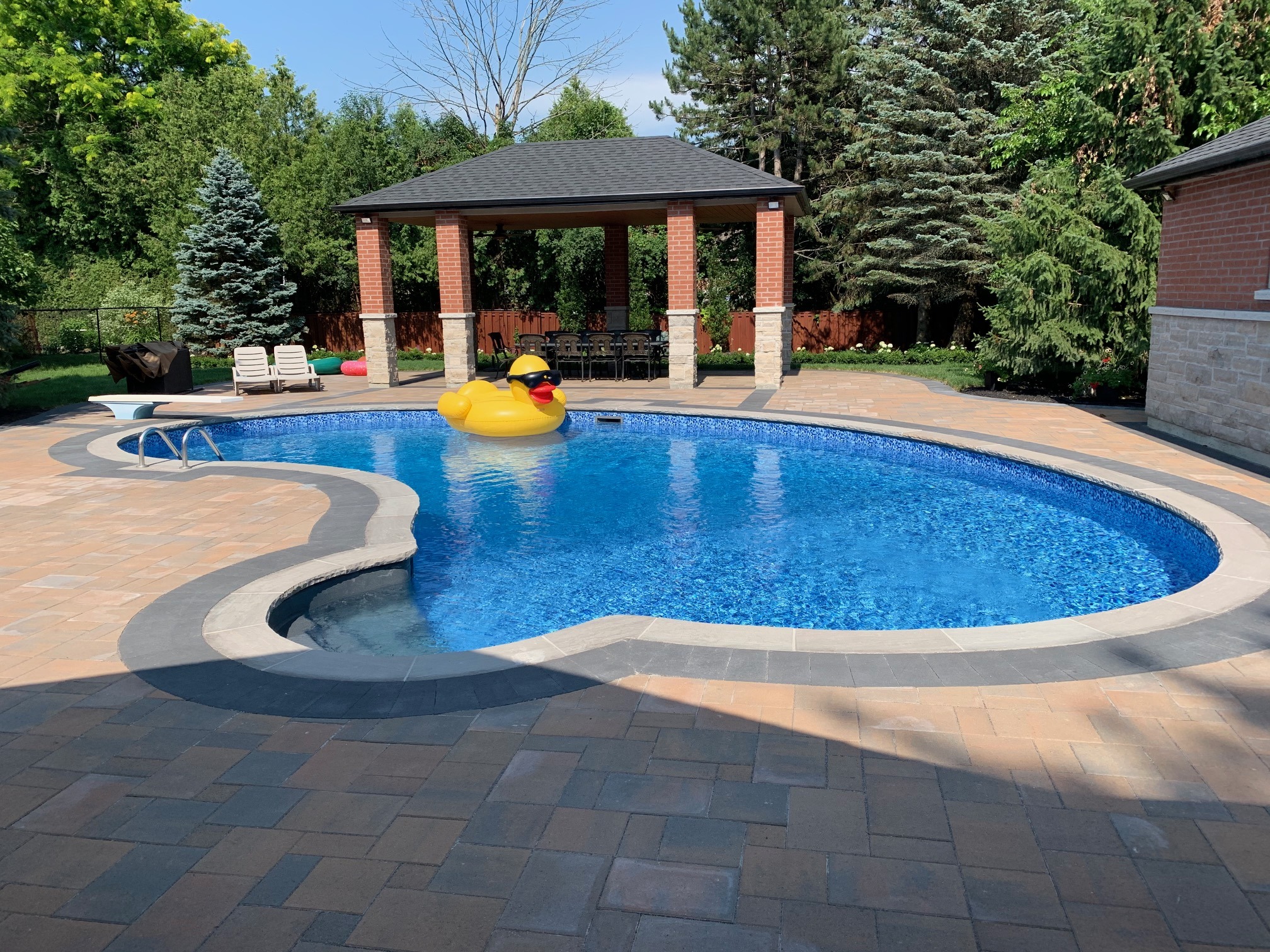 A backyard pool features a large inflatable duck. Nearby is a shaded gazebo, surrounded by trees and patio seating.