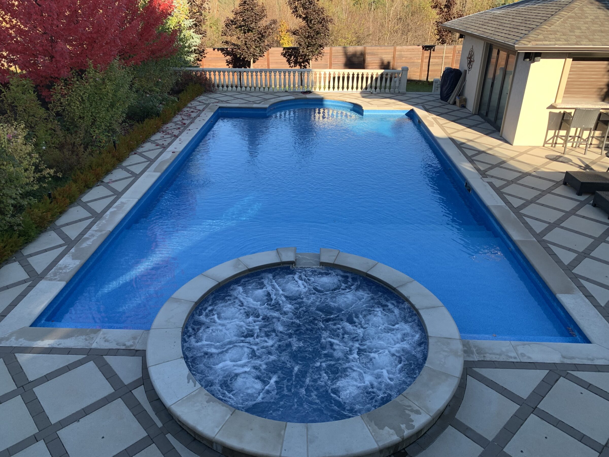 A backyard view with a large rectangular pool, adjacent hot tub, patio area, and vibrant autumn trees surrounding the space.