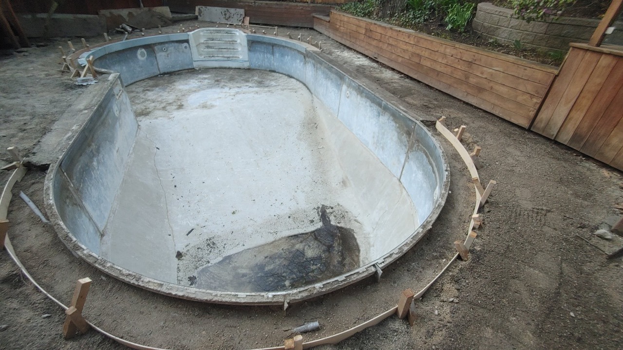 An empty, oval-shaped pool undergoing renovation, surrounded by dirt and wooden planks, with terraced garden walls in the background.