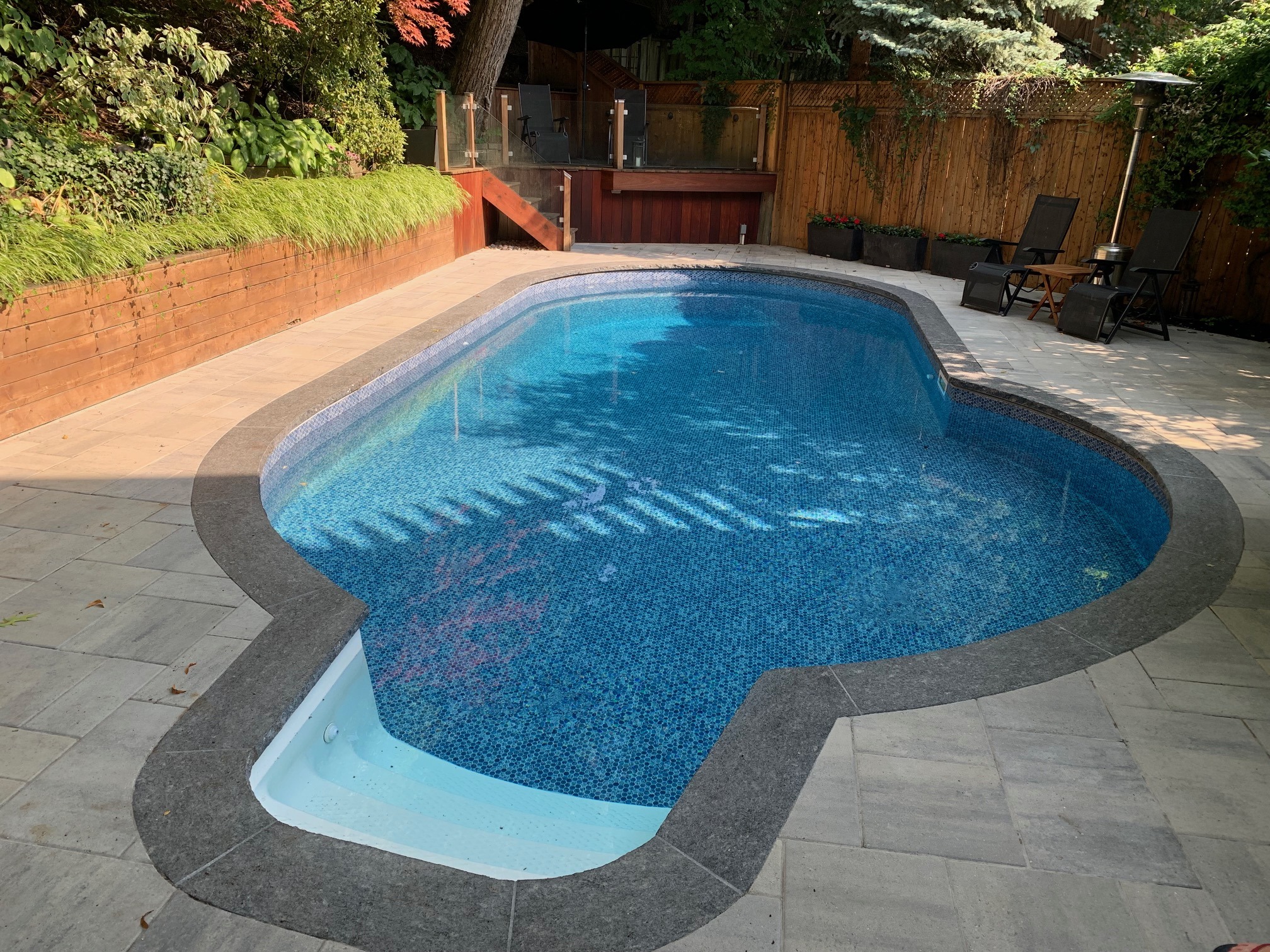 A backyard swimming pool with paving stones, surrounded by lush greenery and wooden fencing, featuring lounge chairs and a small deck area.