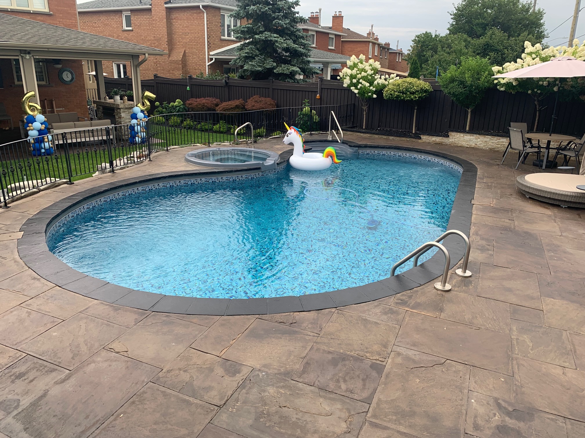 A backyard pool with a unicorn float, surrounded by patio, balloons, and outdoor seating. Brick houses and greenery visible in the background.