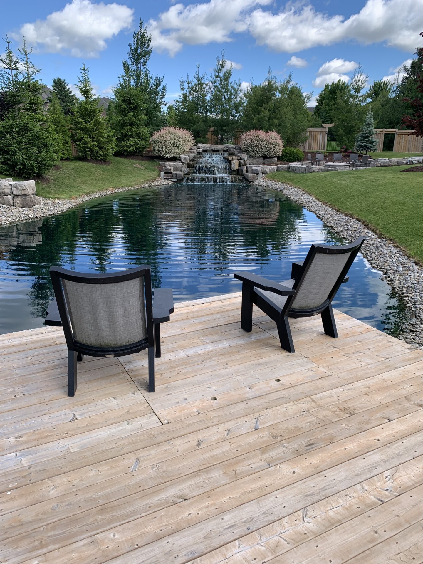Two empty chairs on a wooden deck overlook a tranquil pond with a small waterfall, surrounded by lush greenery under a blue sky.