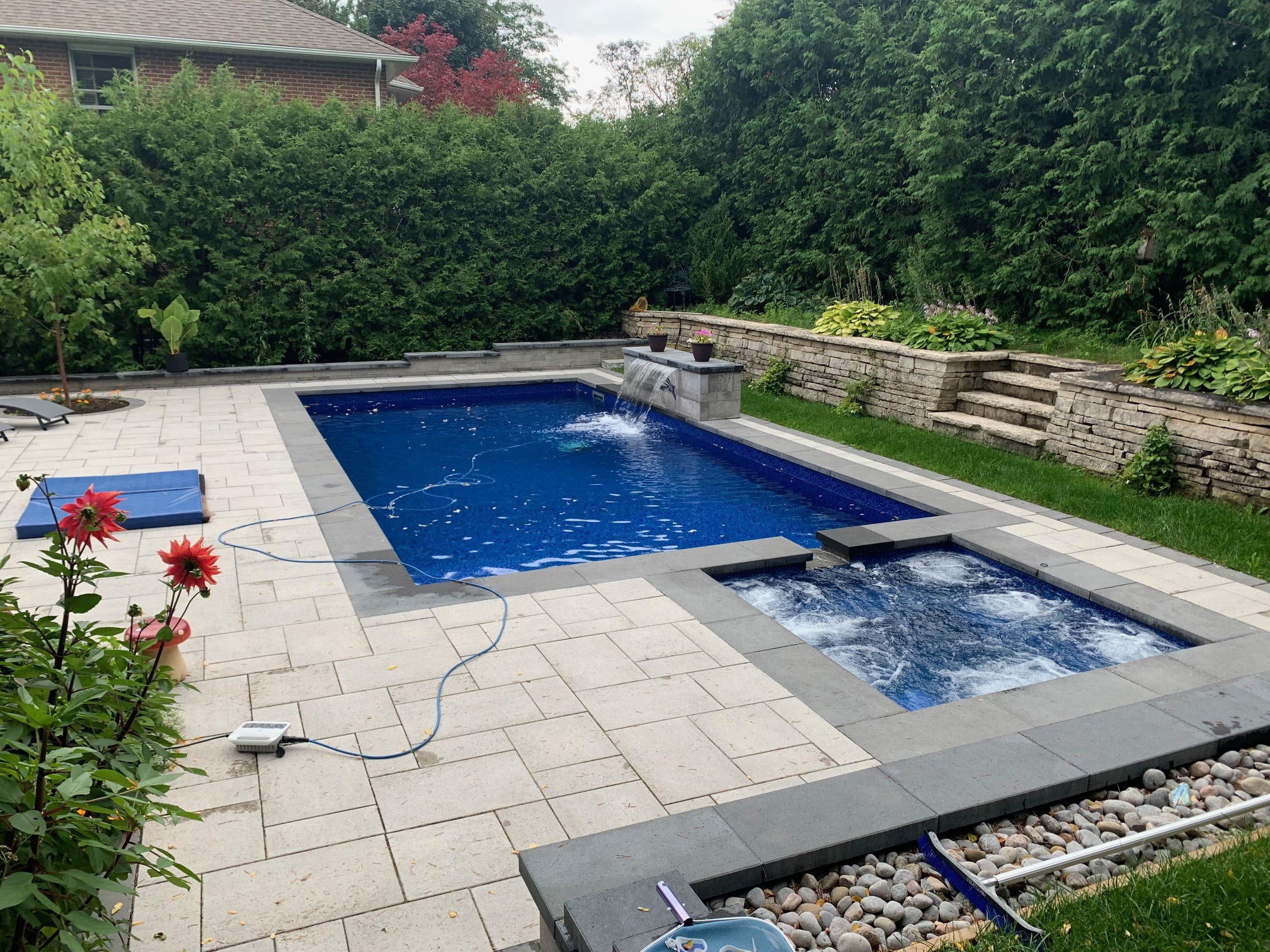 A backyard features a modern swimming pool, surrounded by lush greenery, stone patio, and a cozy seating area near colorful flowers.