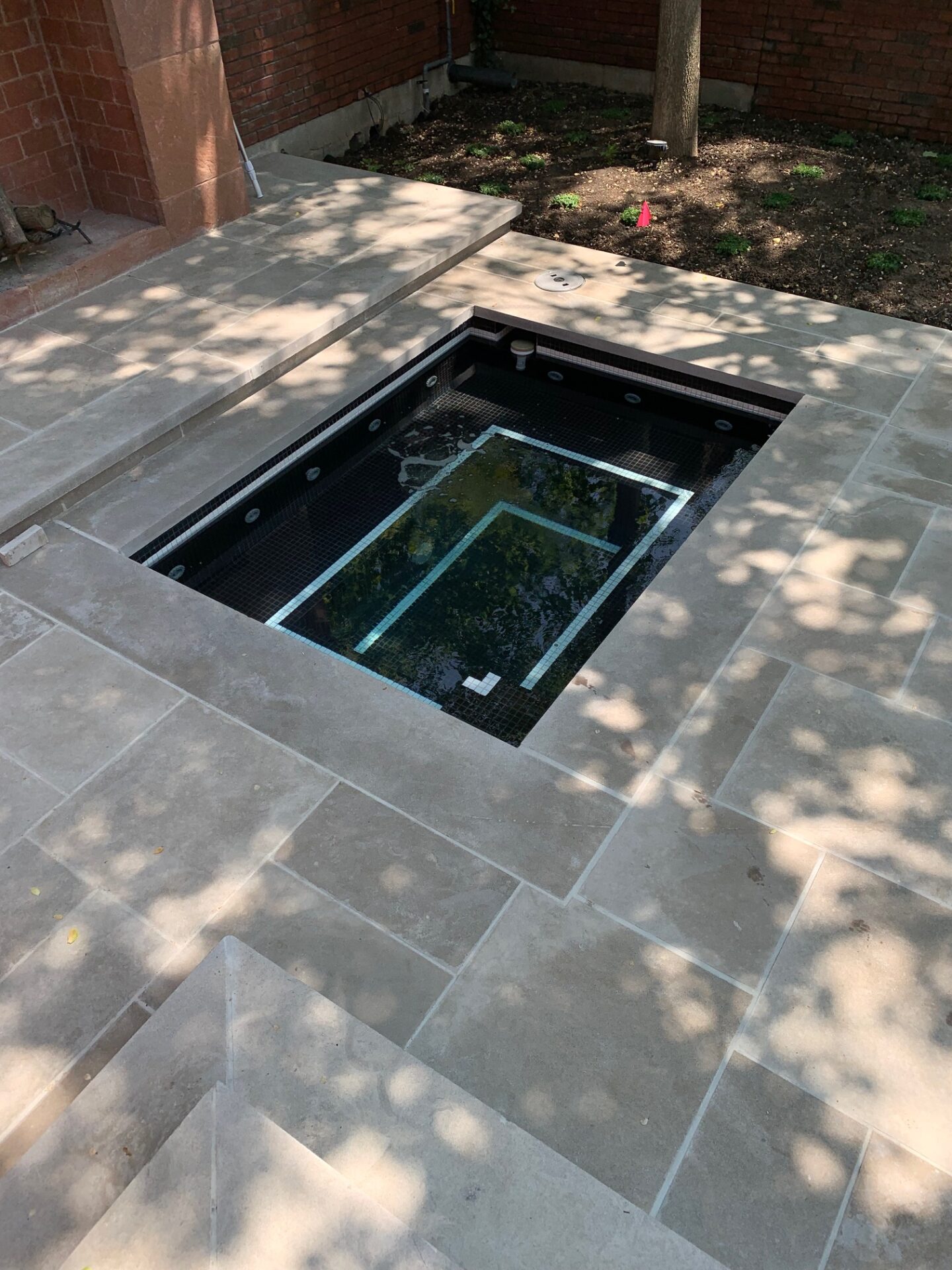 A small, modern, rectangular plunge pool set in a stone-tiled patio area with surrounding red brick walls and shaded landscaping.