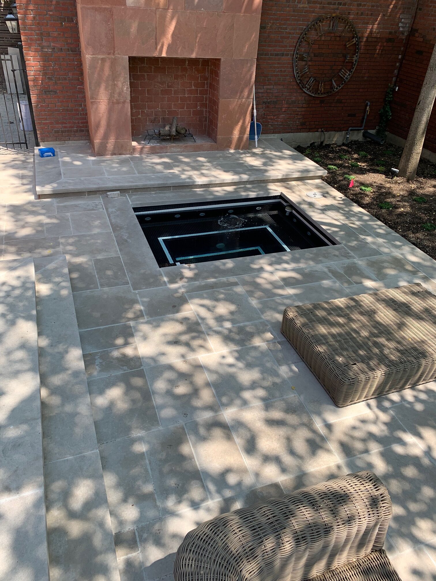 Small patio with fireplace, hot tub, wicker seats, and large clock on brick wall. Dappled sunlight creates patterns on tiled flooring.