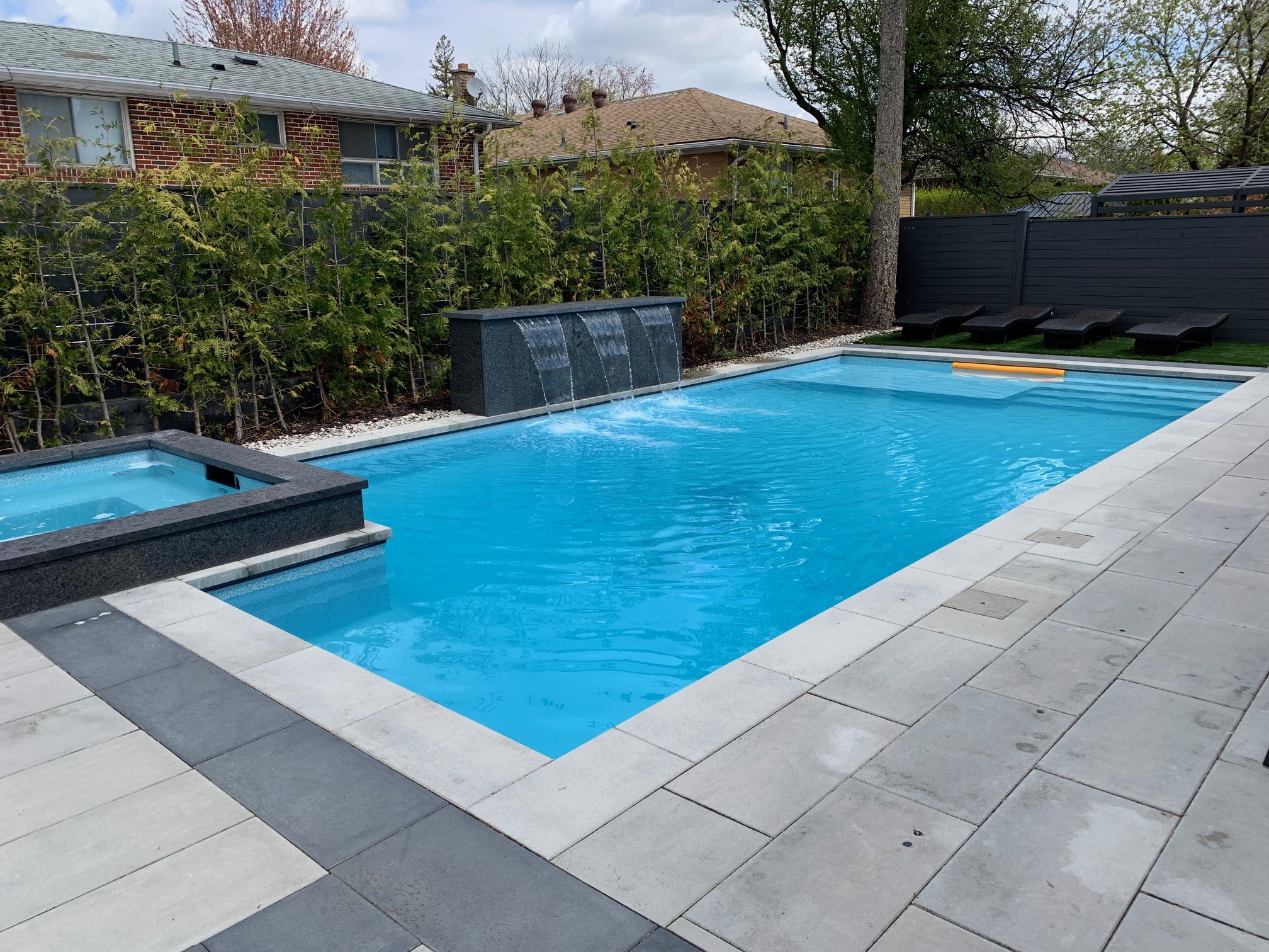 A backyard swimming pool with a water feature, surrounded by patio tiles, trees, lounge chairs, and adjacent to residential houses.