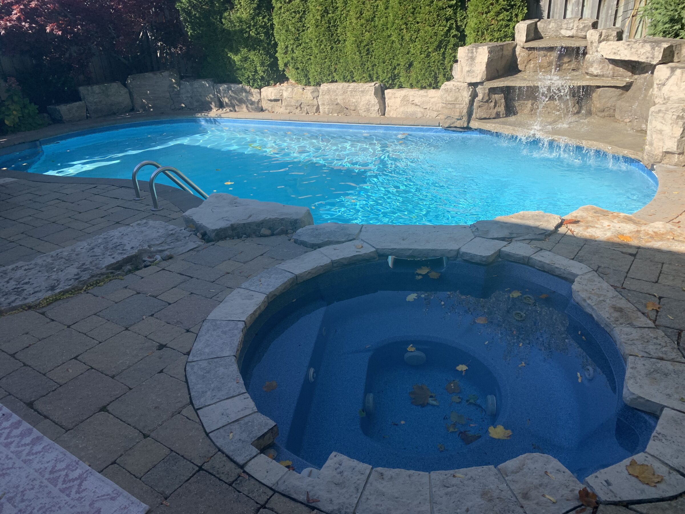 A stone patio surrounds a swimming pool with a waterfall and a connected spa, enclosed by lush greenery and rock features.