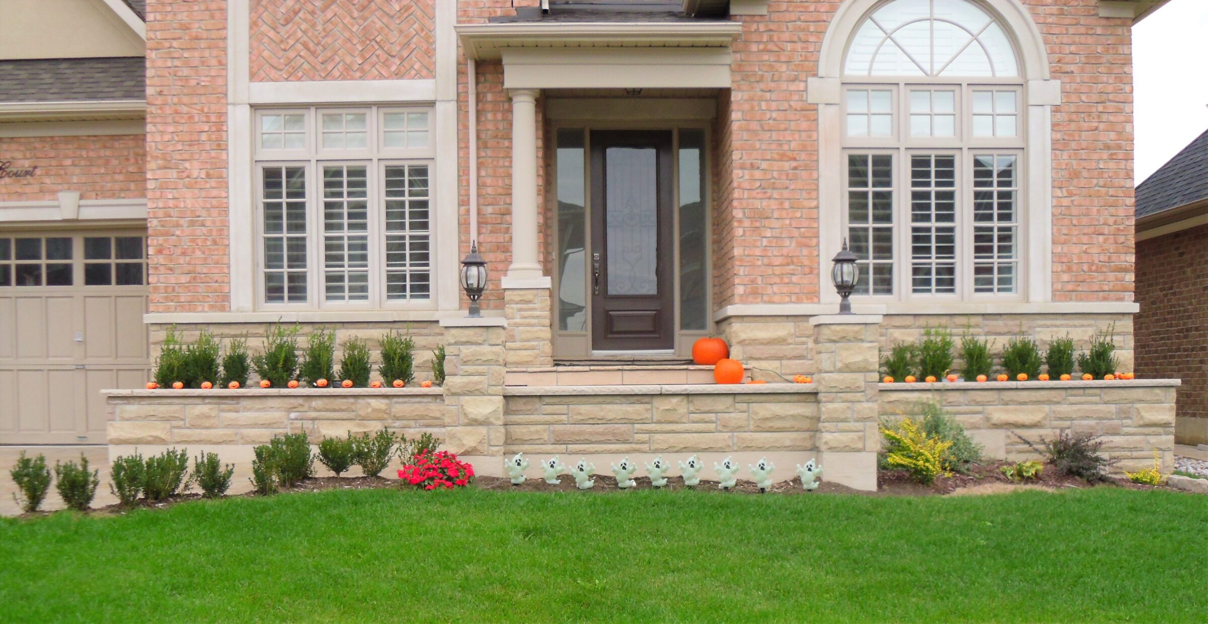 A brick house with a decorated porch featuring pumpkins and faux ghost figures, surrounded by neatly trimmed green shrubs and lawn.