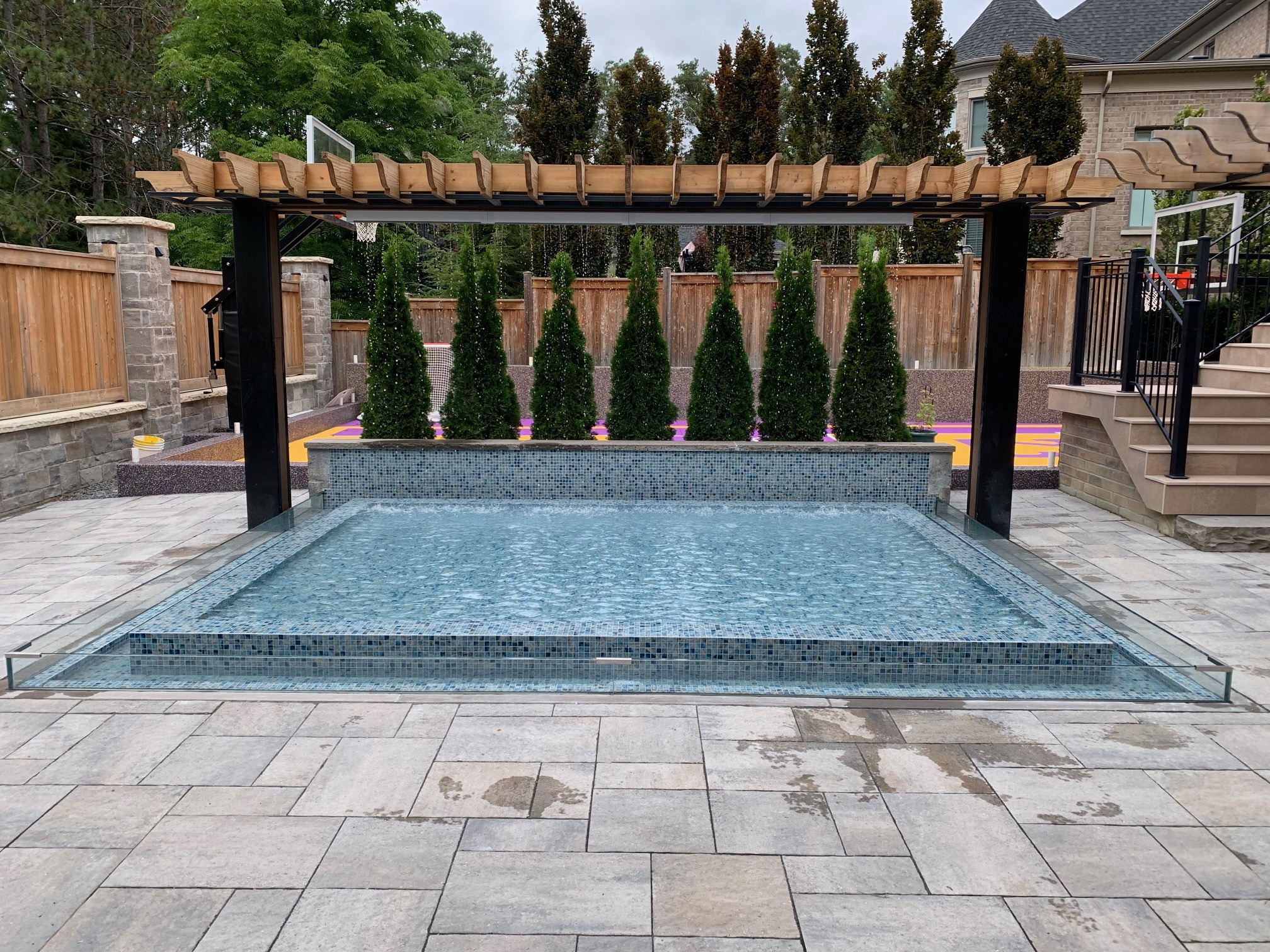 Modern backyard with a small reflective pool, pergola, neatly trimmed trees, paved patio, and a glimpse of a colorful basketball court in the background.
