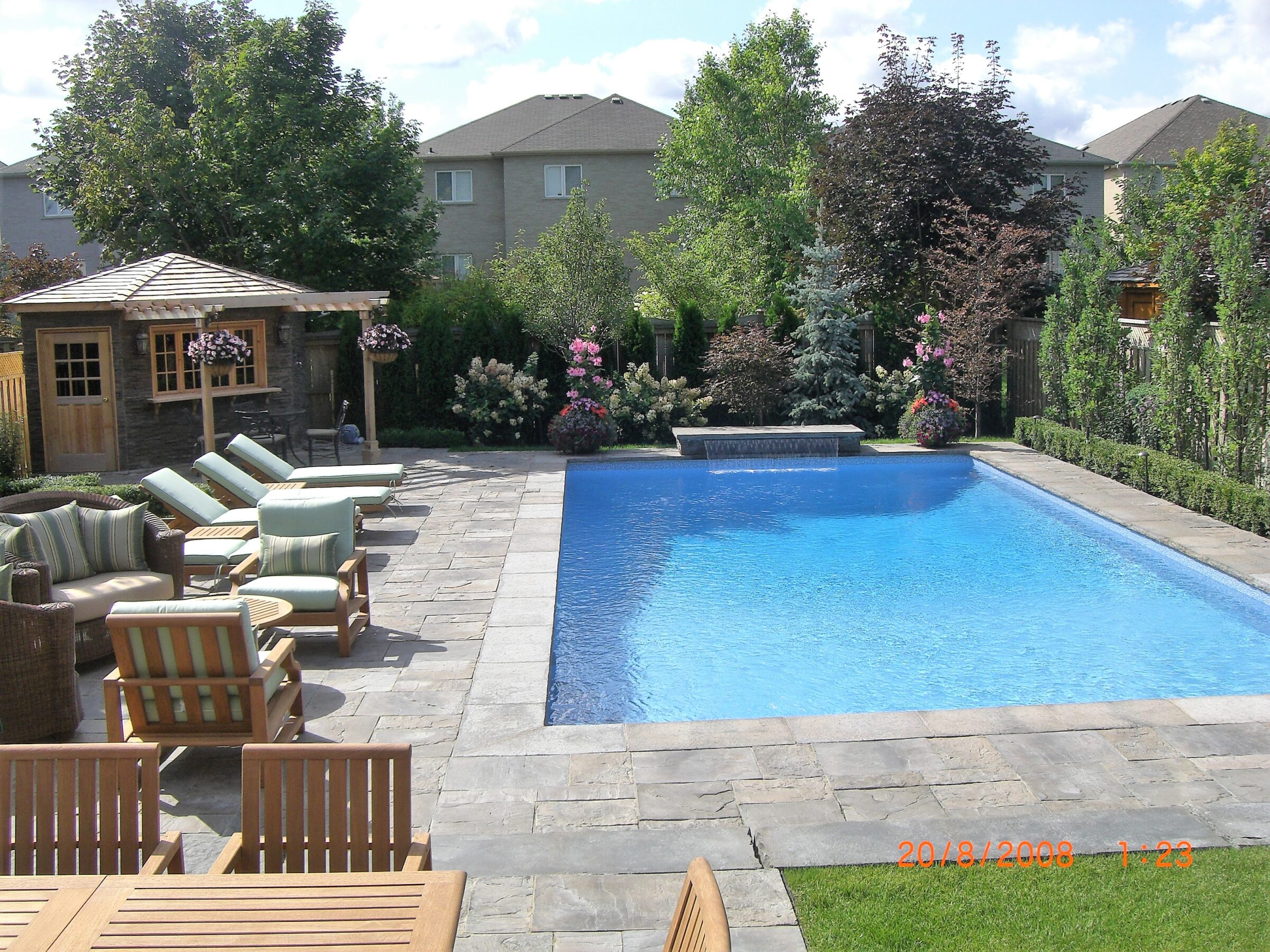 Sunny backyard with blue pool, cushioned loungers, and wooden pergola. Surrounded by well-maintained trees and bushes, creating a serene, private atmosphere.
