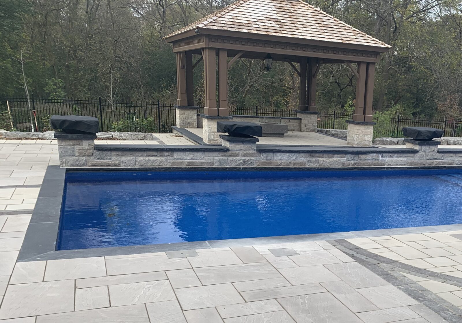 A serene backyard features a blue swimming pool, stone patio, and wooden gazebo, surrounded by trees and a metal fence.