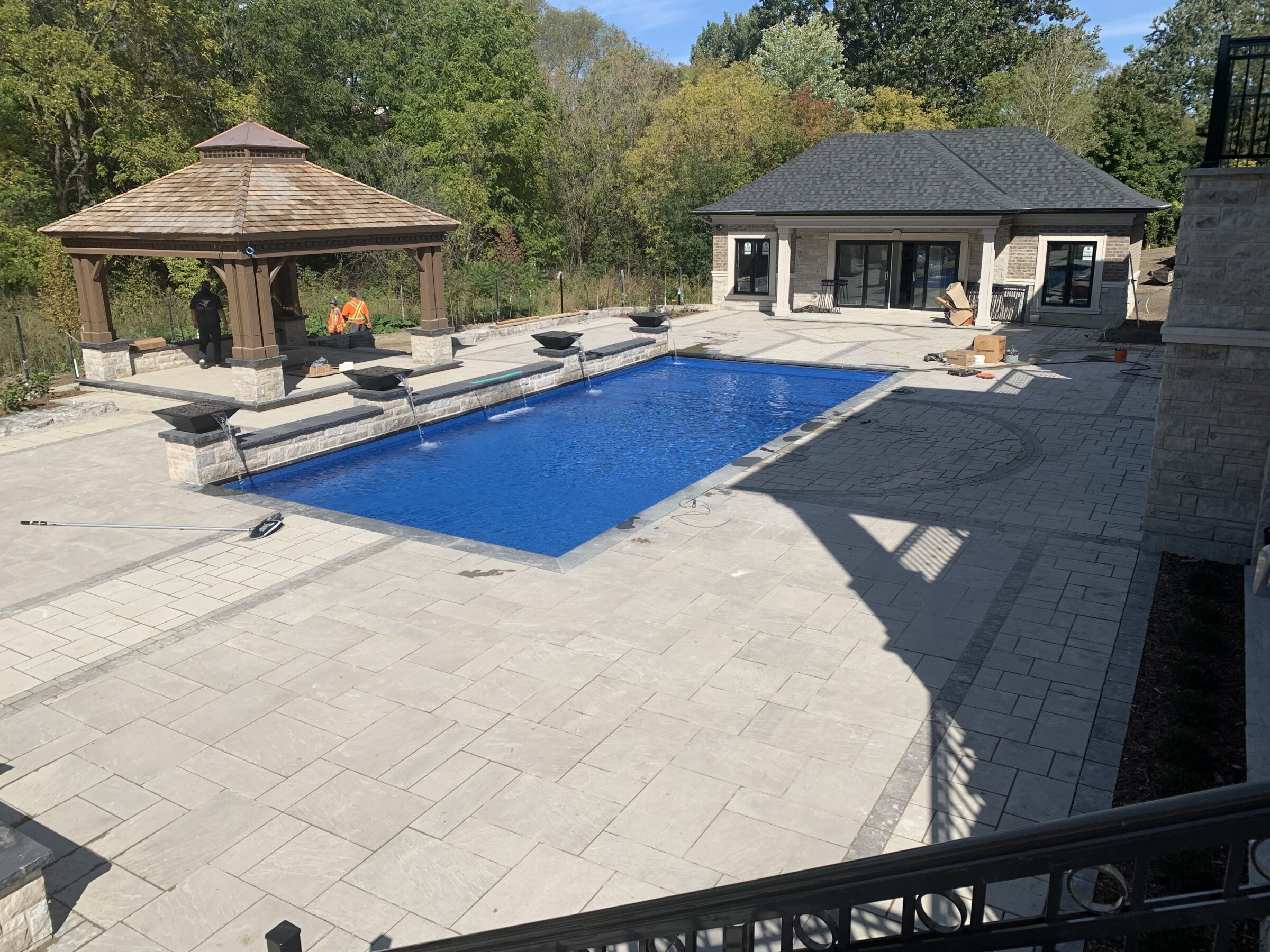 A modern backyard pool with a gazebo, two people nearby, surrounded by trees, and a small building in the background.
