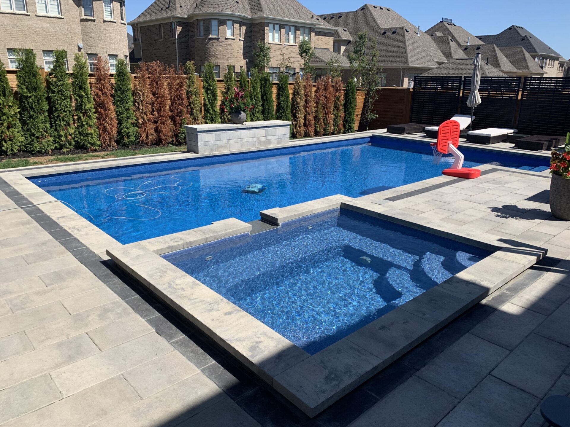 A residential backyard with a modern pool and hot tub, surrounded by brick houses and tall plants, under clear blue skies.
