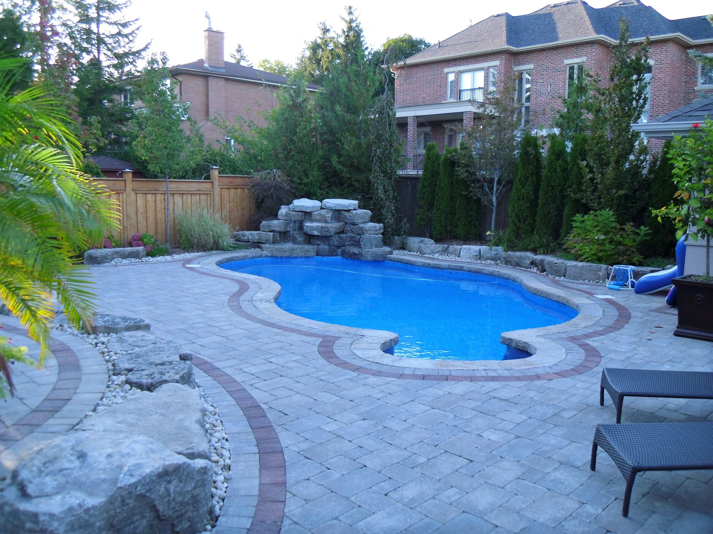 A backyard with a kidney-shaped pool, stone waterfall feature, lounge chairs, brick-paved patio, surrounded by trees, plants, and wooden fence.
