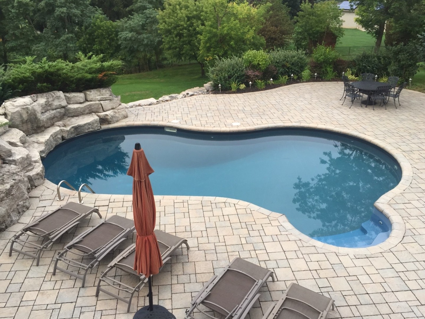 A backyard features a curved pool, lounge chairs, a closed umbrella, and patio table surrounded by greenery and stone landscaping.