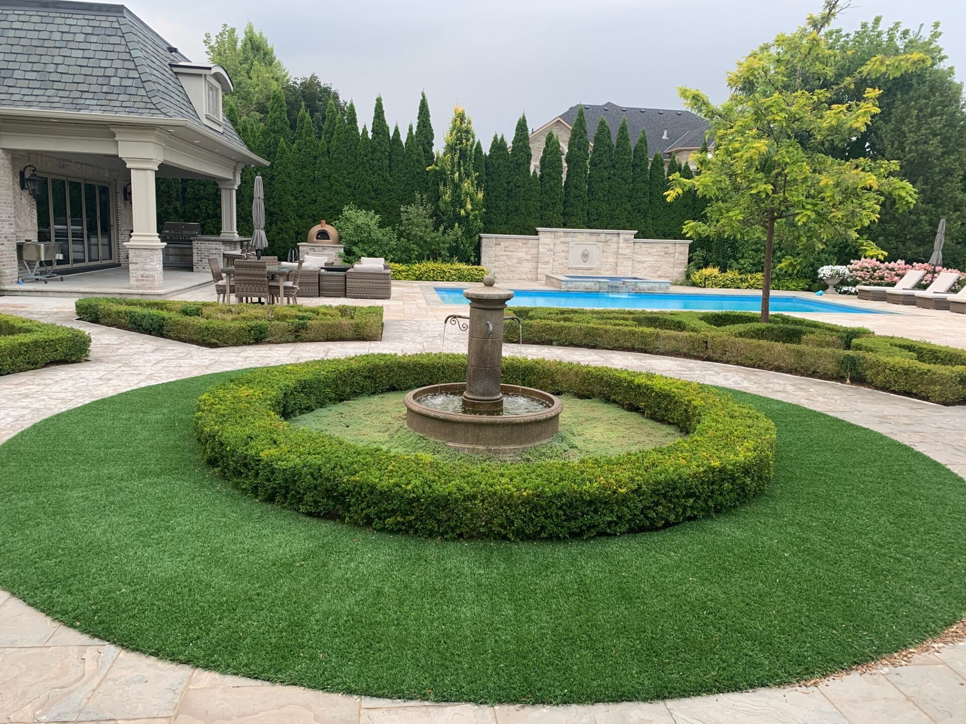 Elegant garden with a central fountain, lush hedges, patio furniture, and a swimming pool. Trees surround the area, creating a serene atmosphere.