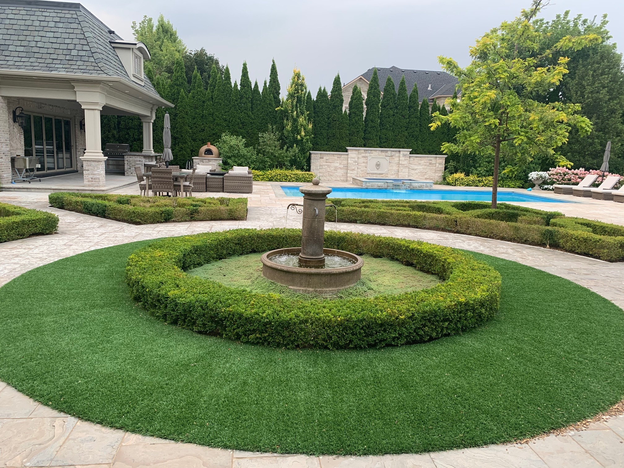 Elegant backyard features a circular fountain, lush greenery, a swimming pool, outdoor seating, and a shaded patio with a pizza oven.