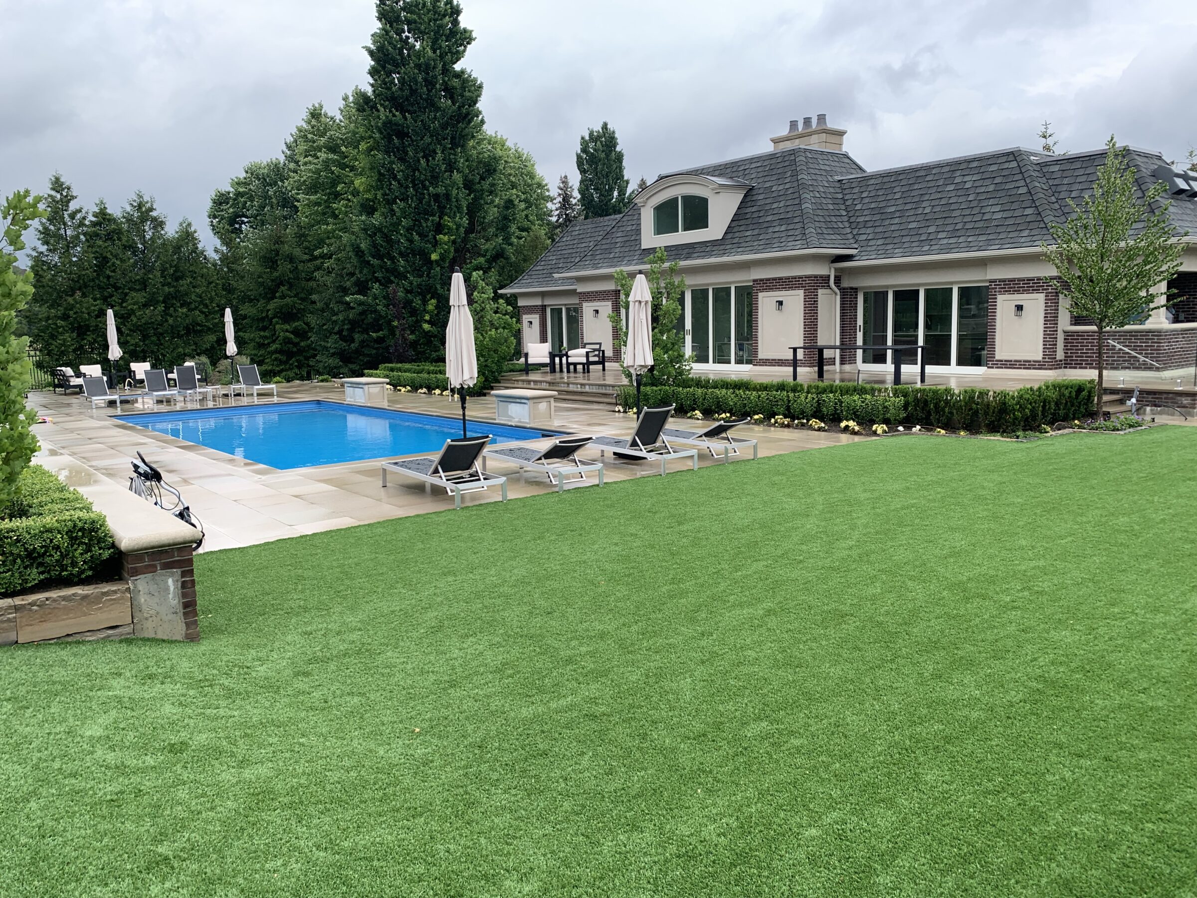 Modern house with a large backyard features a swimming pool, lounge chairs, and neatly trimmed greenery, set against a cloudy sky backdrop.