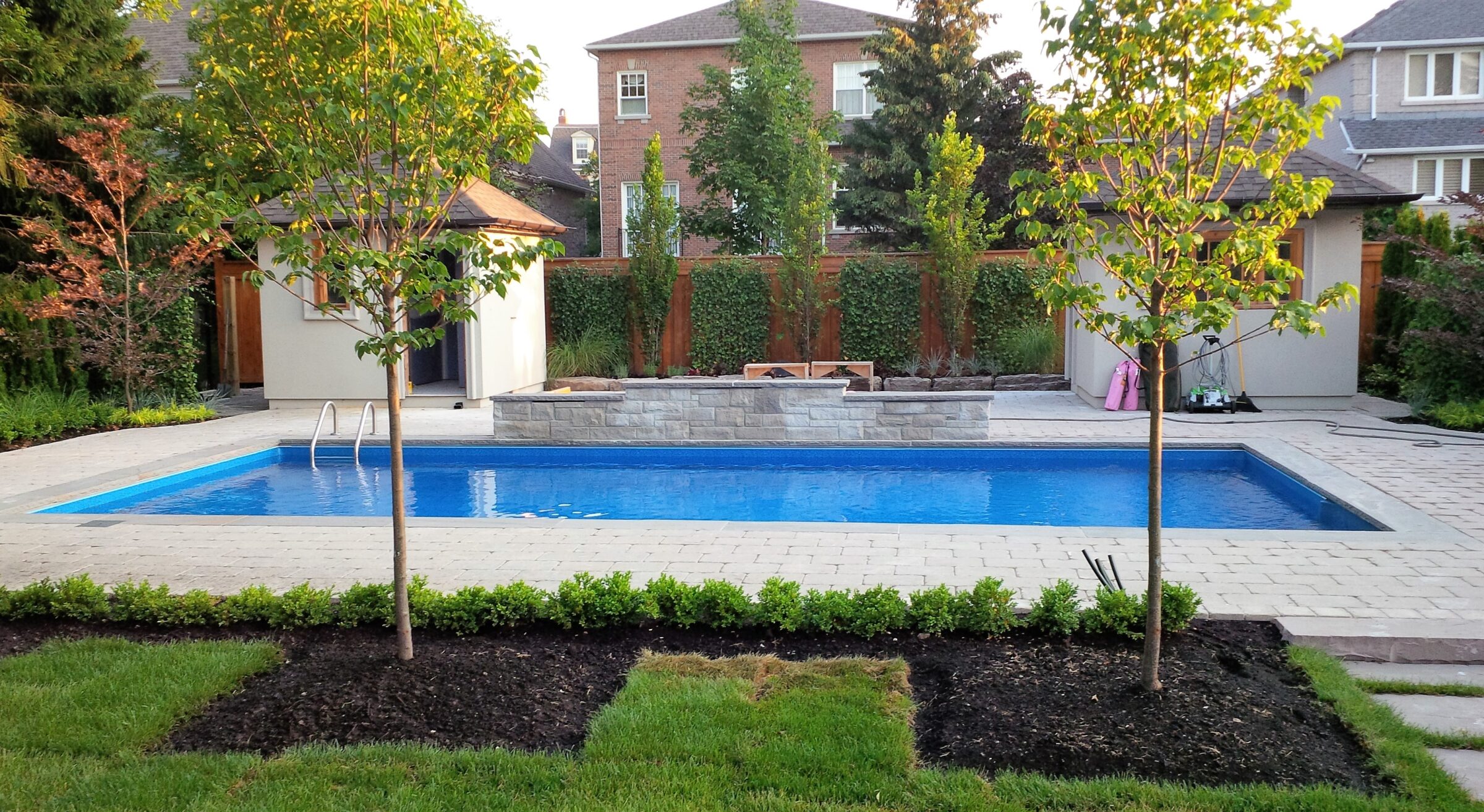 A backyard with a rectangular swimming pool, surrounded by trees, small garden area, stone walkway, and two small buildings or sheds.