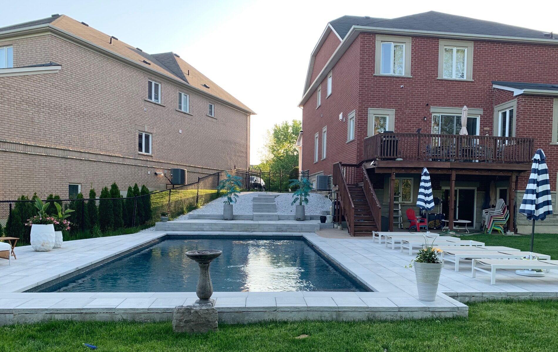 Two brick houses with a backyard pool area, featuring sun loungers, potted plants, and striped umbrellas, surrounded by well-maintained green grass.