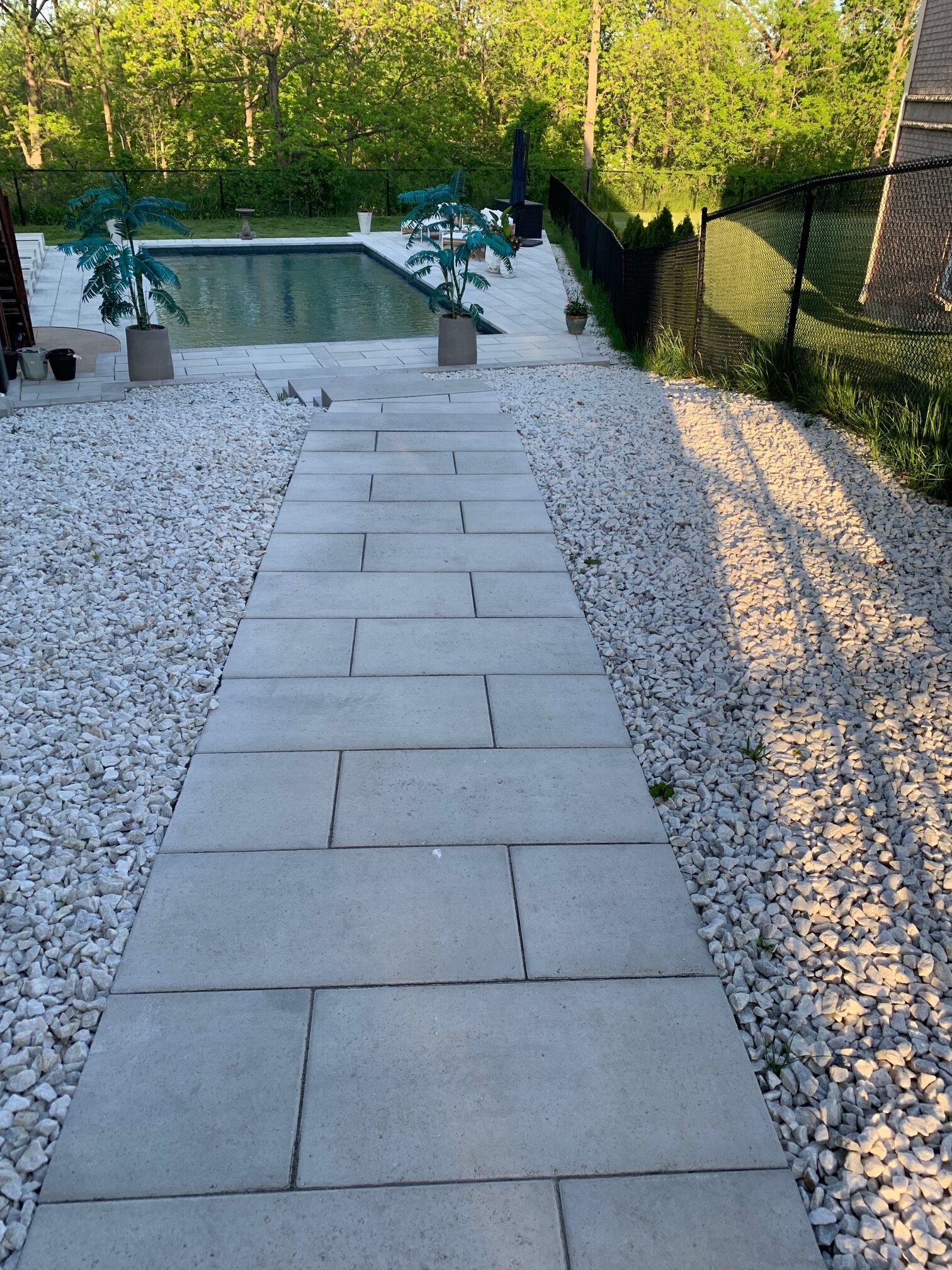 A backyard features a rectangular pool, surrounded by trees and decorative potted plants, with a paved walkway and gravel landscaping.