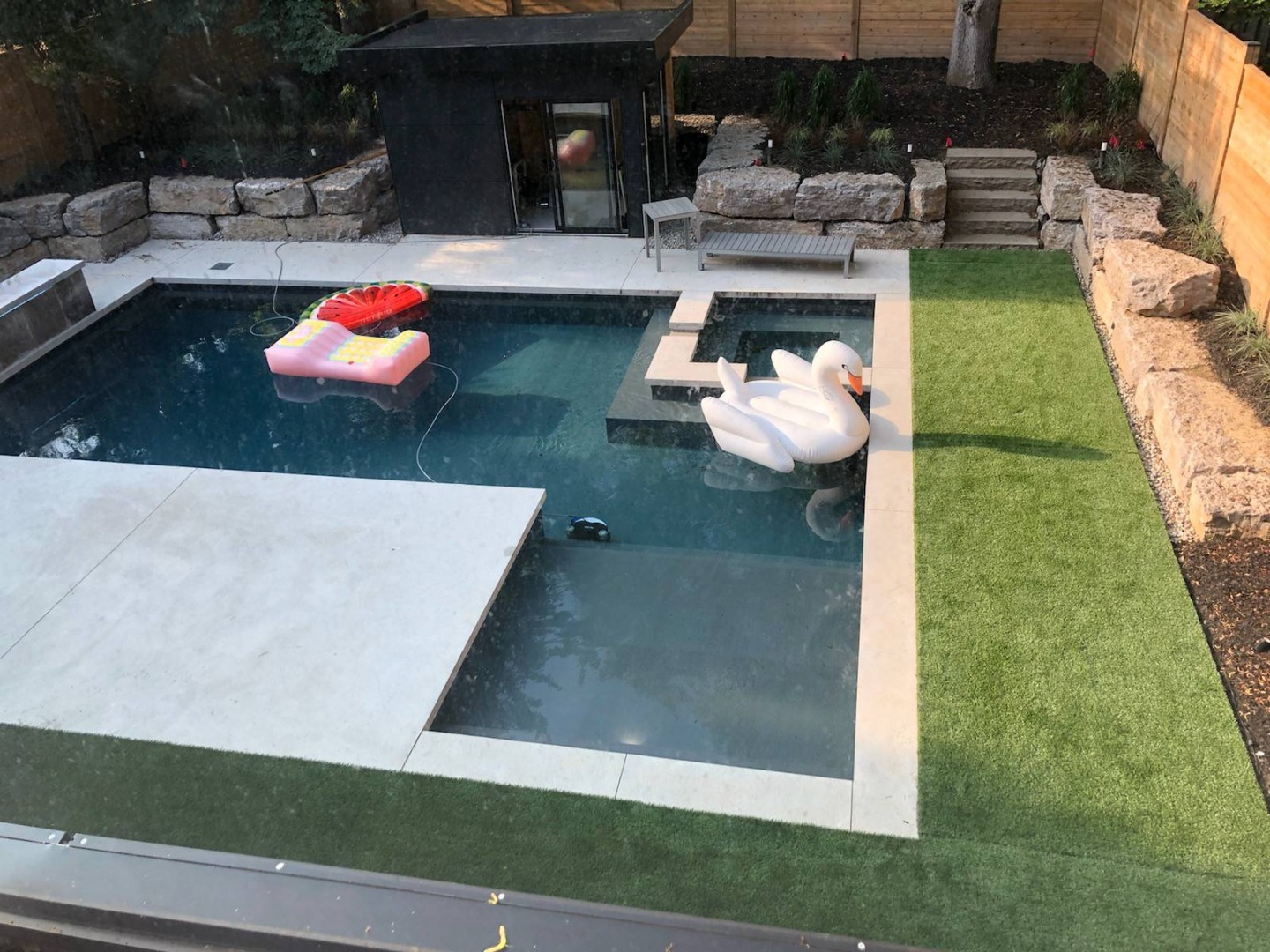 Modern backyard with a pool, inflatable swan, and watermelon float. Surrounded by stone walls, lush greenery, and a small garden shed.