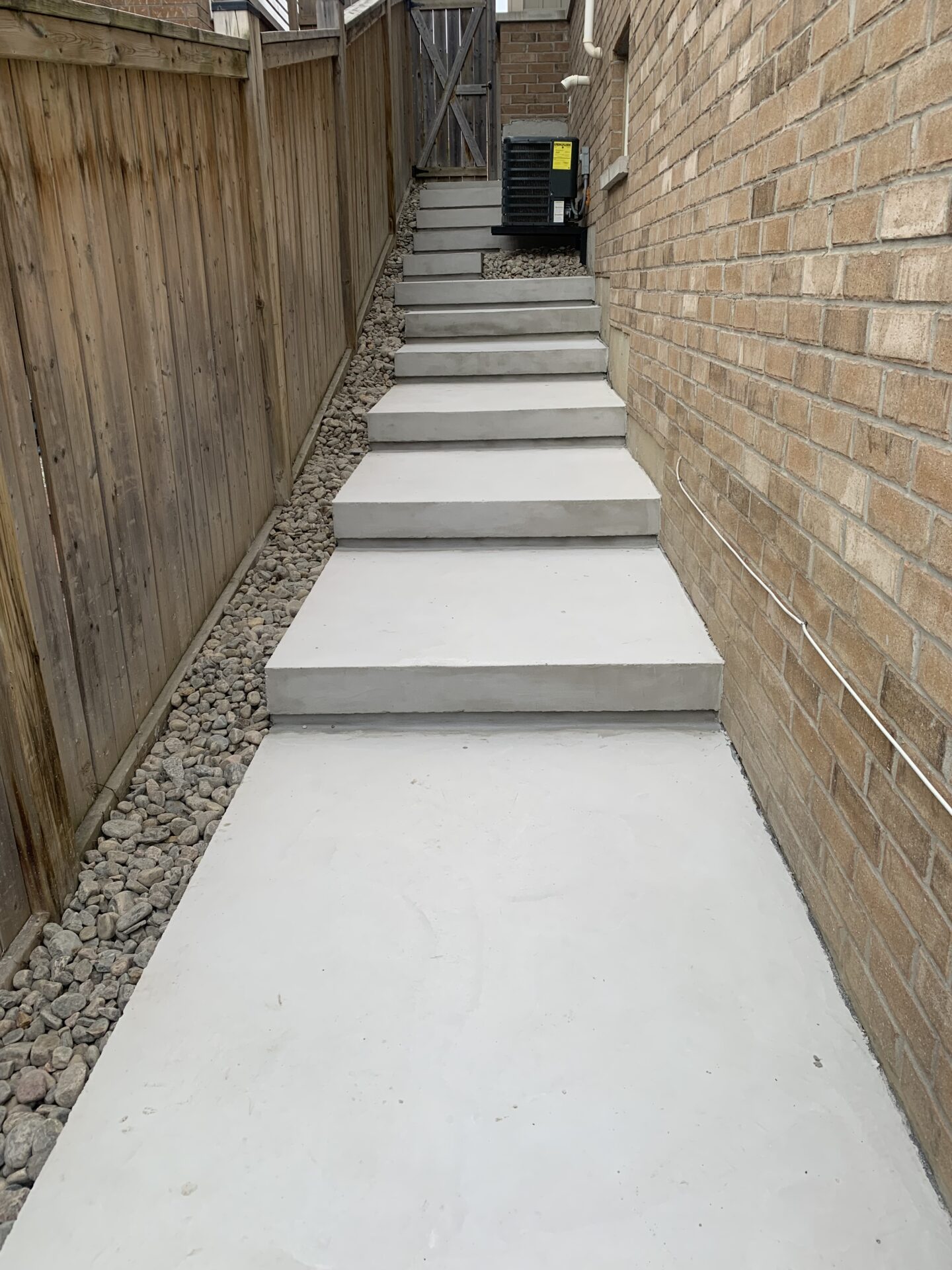 Concrete steps bordered by a wooden fence and brick wall, leading up to a wooden gate with stones alongside the path.