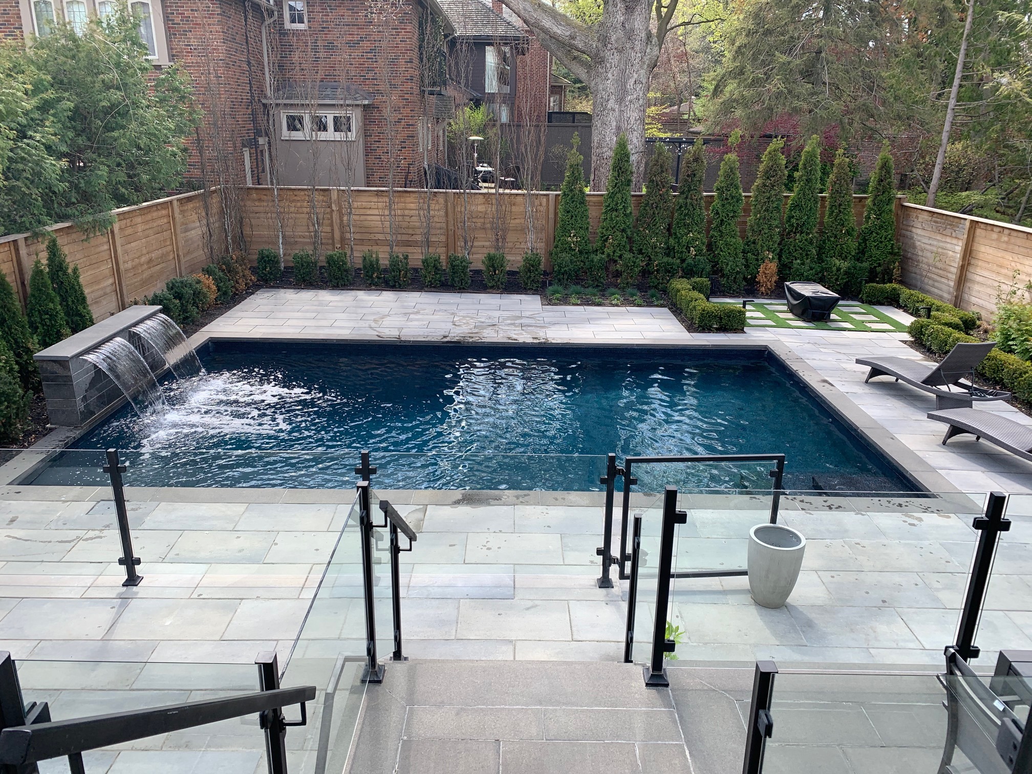 A neatly landscaped backyard features a rectangular pool with a waterfall, surrounded by stone tiles, chairs, and lush greenery.