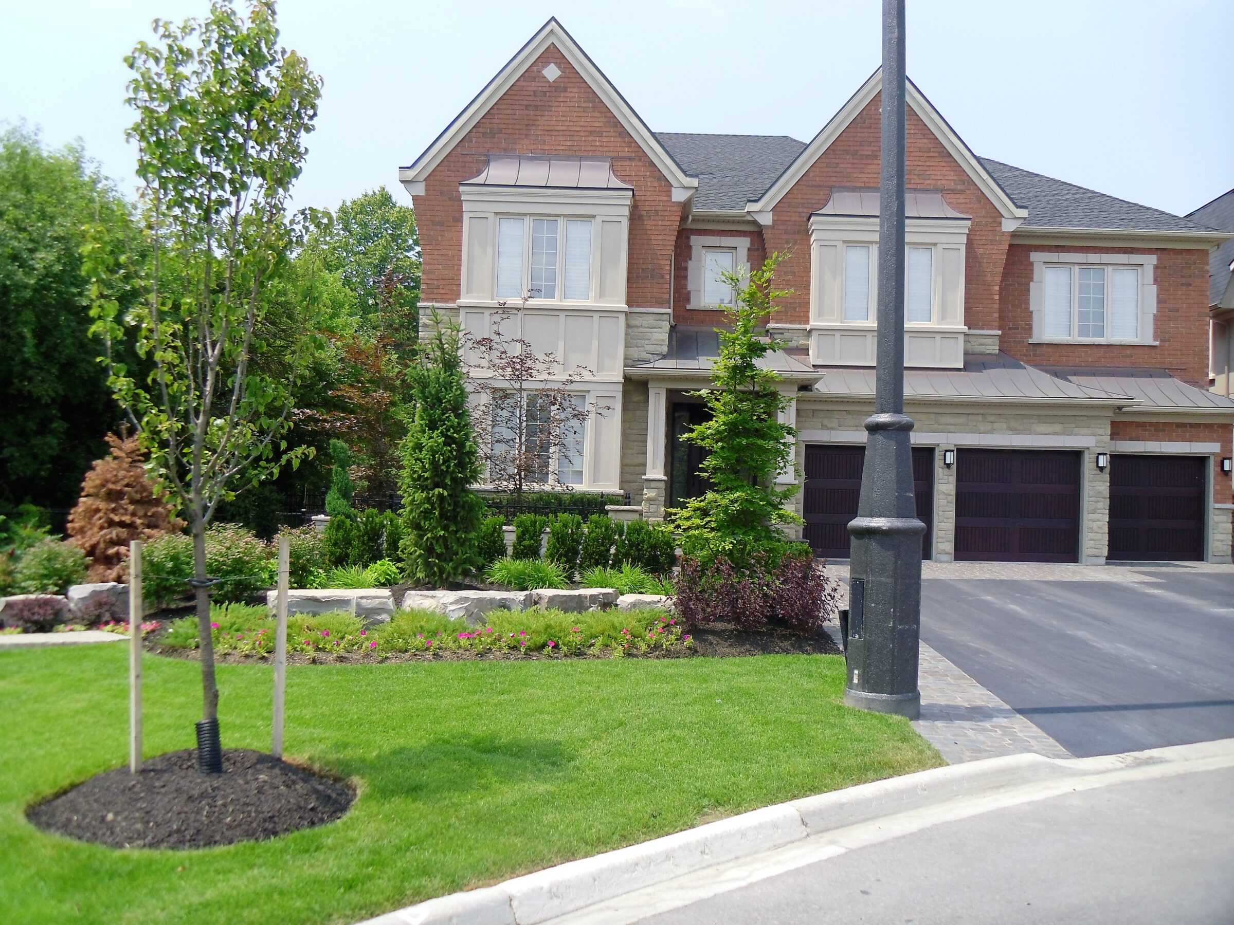 A suburban house with a manicured lawn, young trees, and a lamppost. The brick structure features multiple garage doors and large windows.