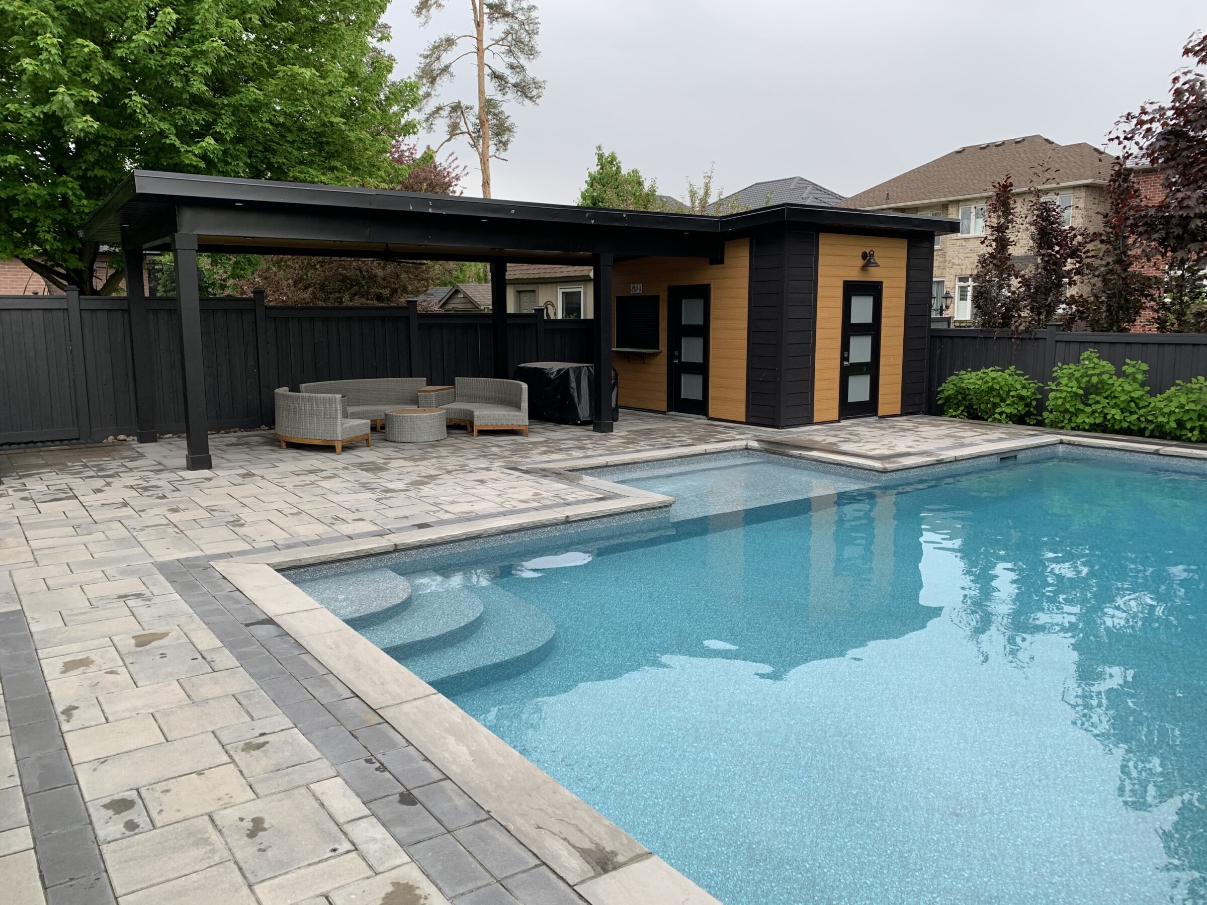 A modern backyard features a pristine swimming pool, patio furniture under a pergola, and a small wooden pool house, surrounded by trees.