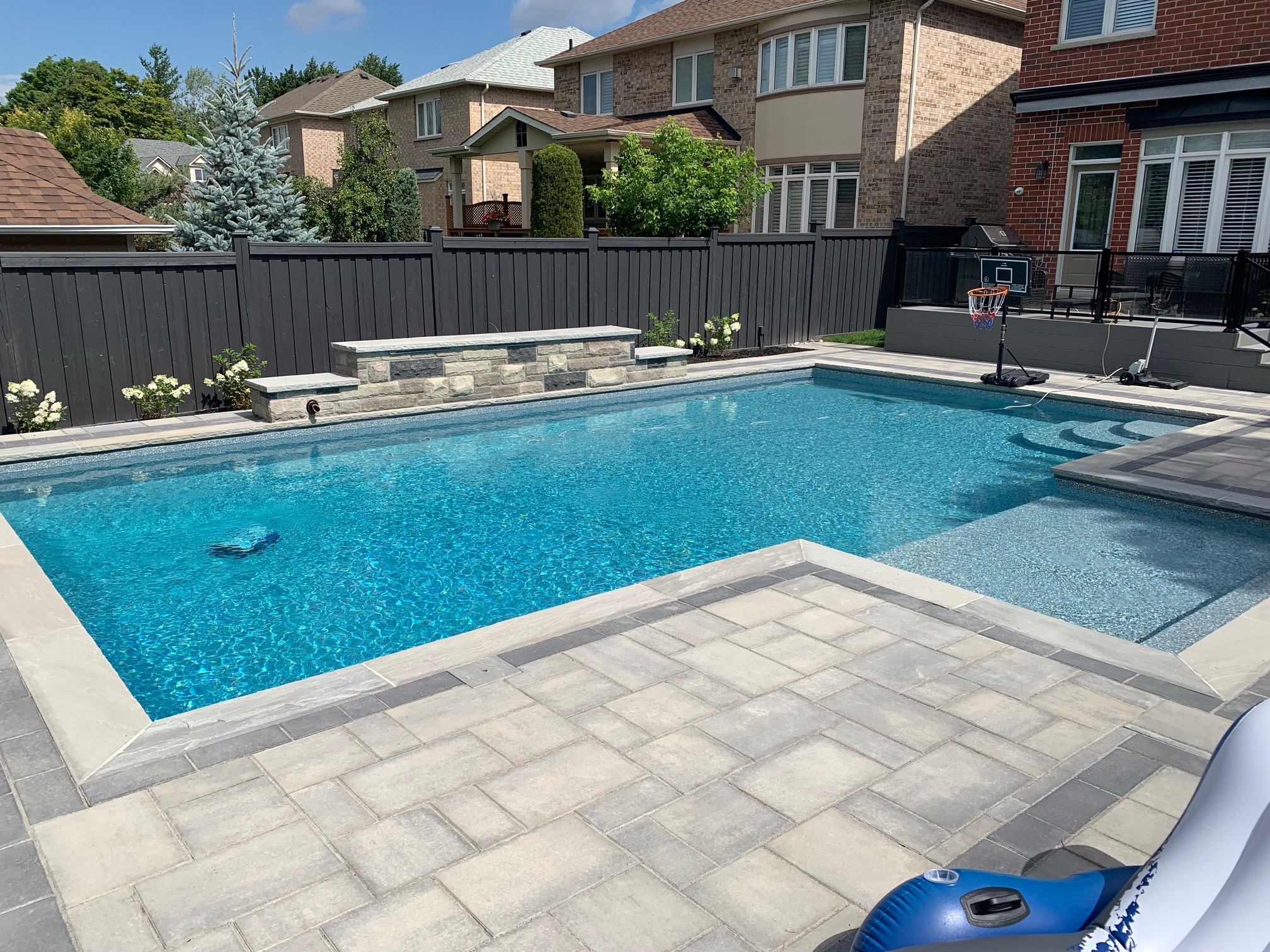A backyard with a clear blue swimming pool, surrounded by patio tiles, privacy fence, houses, and a small basketball hoop setup.