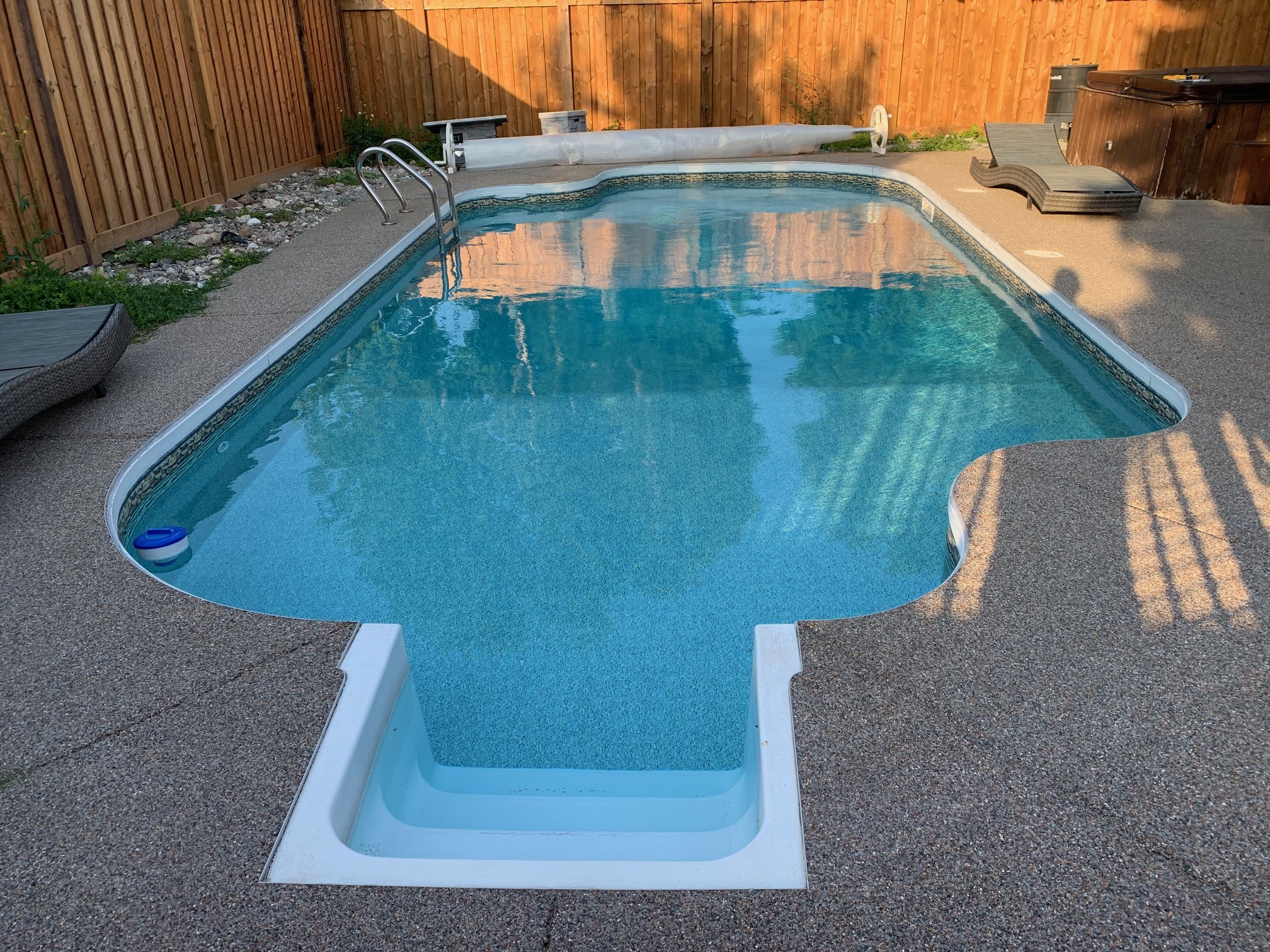 A backyard scene with a clean swimming pool, two lounge chairs, and wooden fencing, under clear daylight conditions. No people present.
