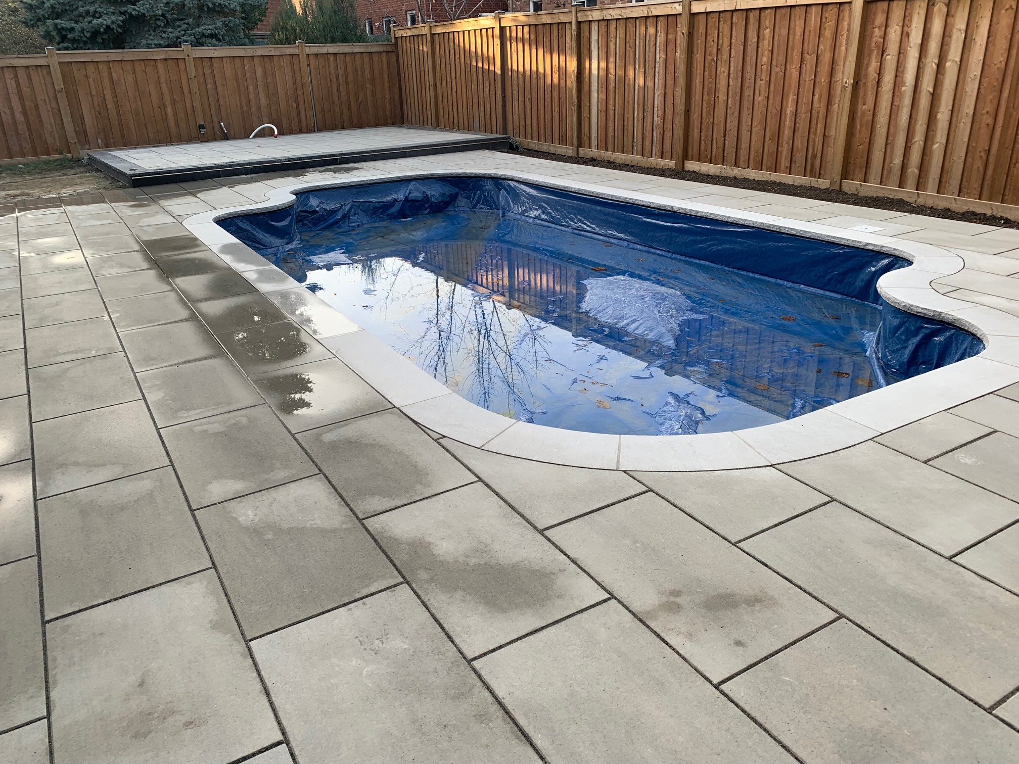 A backyard with a kidney-shaped swimming pool, surrounded by stone tiles, enclosed by a wooden fence, with trees reflected in the water.