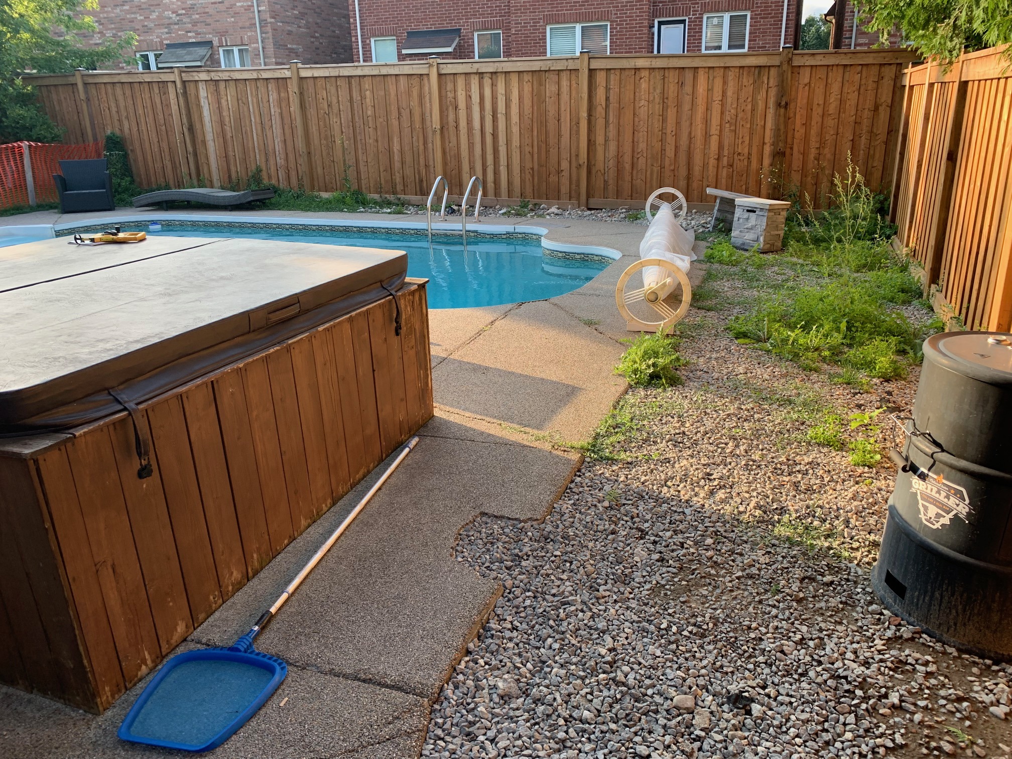 A backyard with a swimming pool, hot tub, grill, lounge chair, and wooden fence, surrounded by greenery and pebbles.