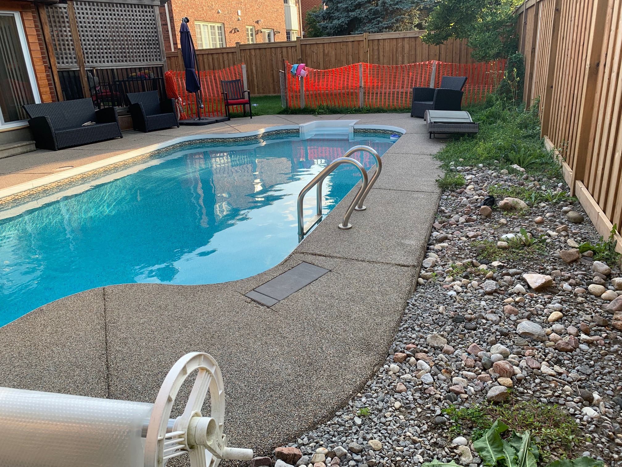 A backyard pool with lounge chairs, gravel landscaping, and a wooden fence. Red mesh fencing and patio furniture are visible.