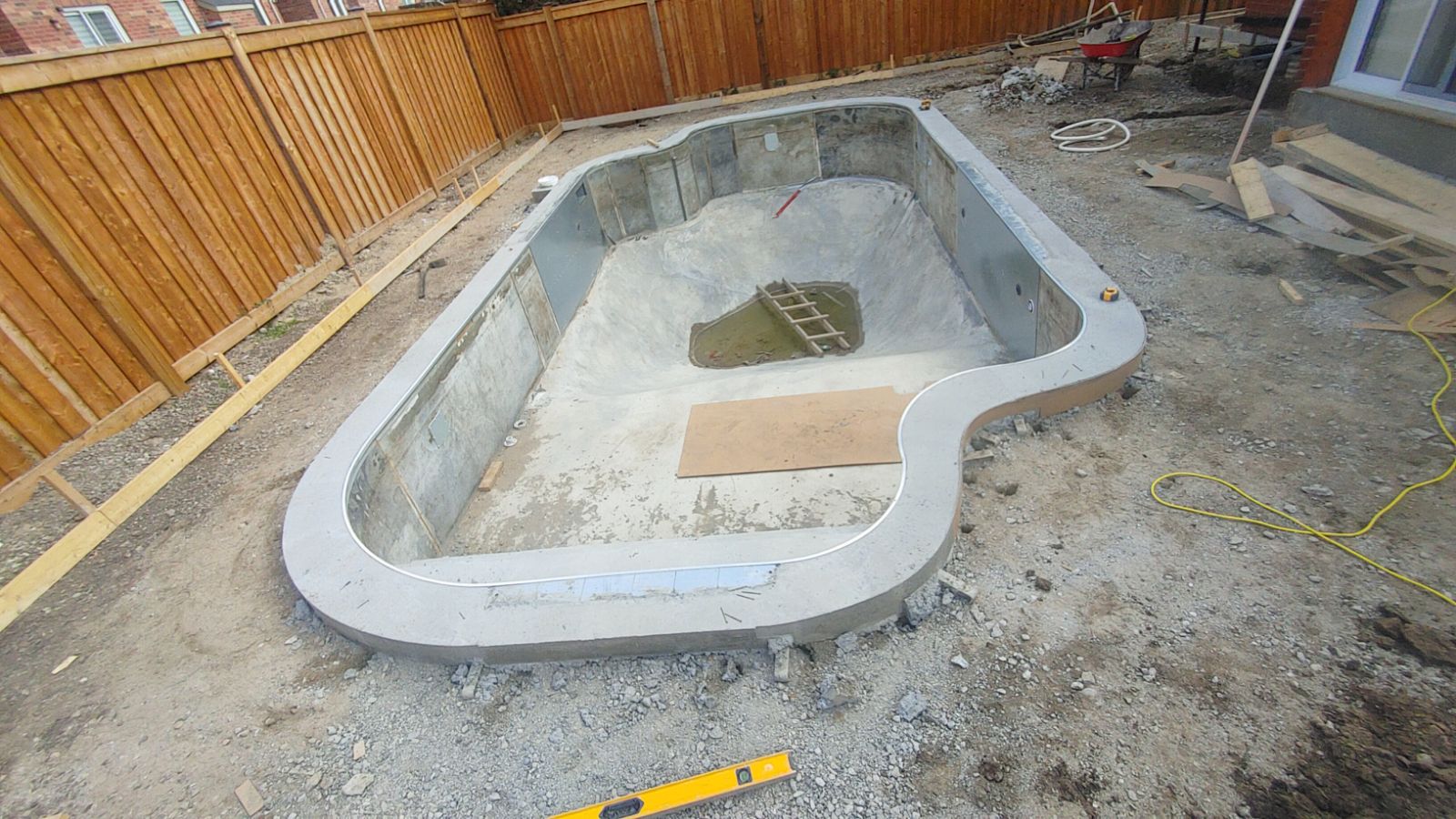 An unfinished swimming pool under construction in a backyard, surrounded by wooden fencing and construction materials scattered around.