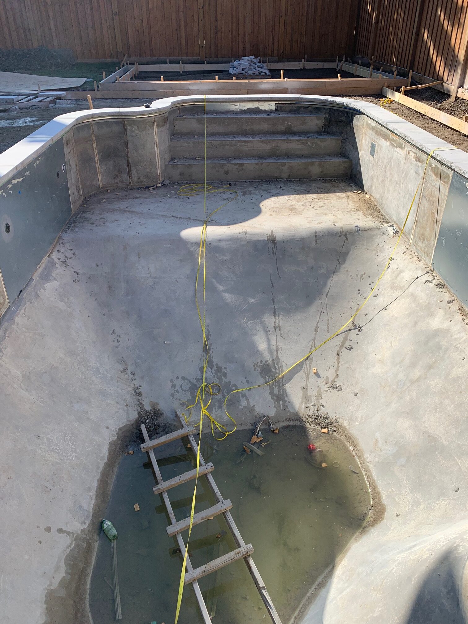 An empty pool under construction with a wooden ladder and water at the bottom, surrounded by a wooden fence and building materials.