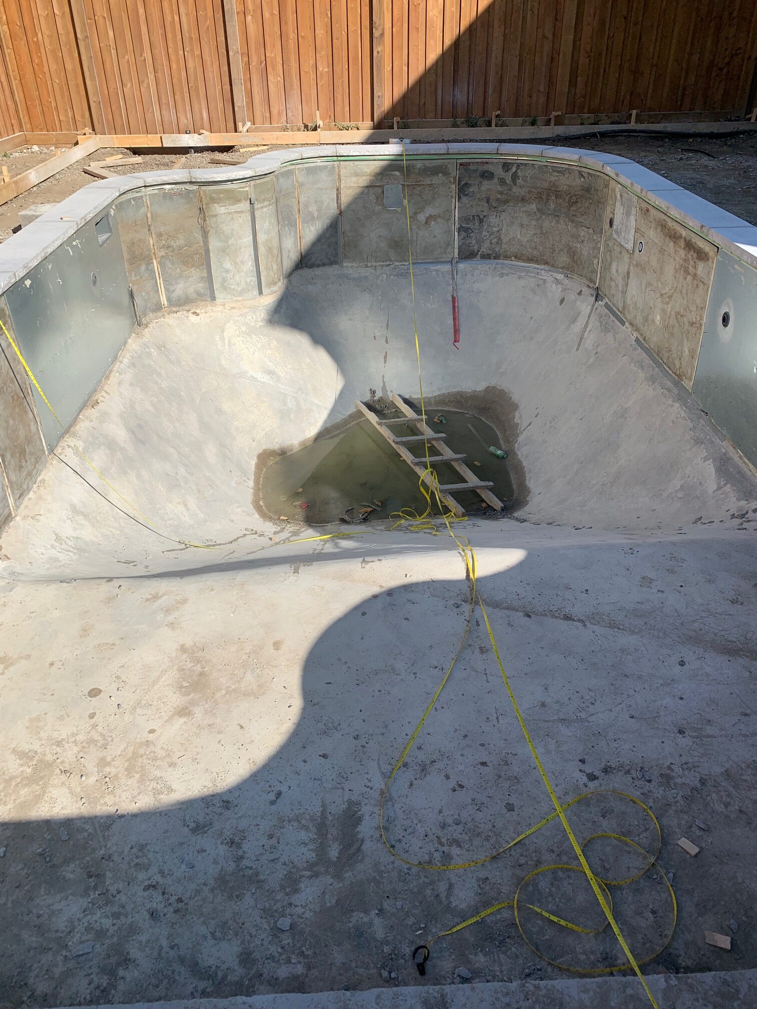 Construction site of an empty pool with a ladder and ropes inside. Wooden fence in the background under sunlight.