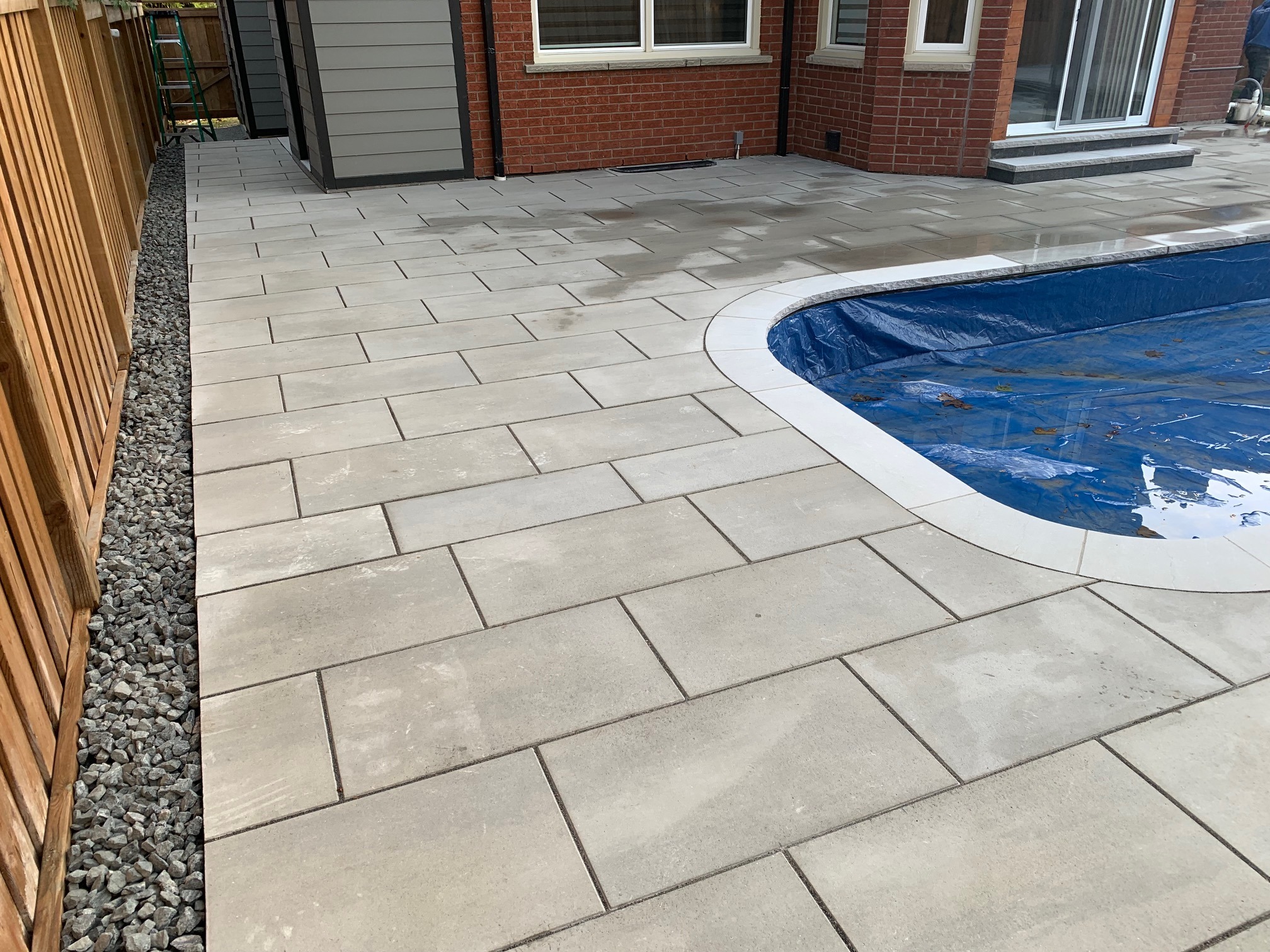 A backyard with a newly paved patio, covered pool, and wooden fence adjacent to a brick house with large windows and sliding door.