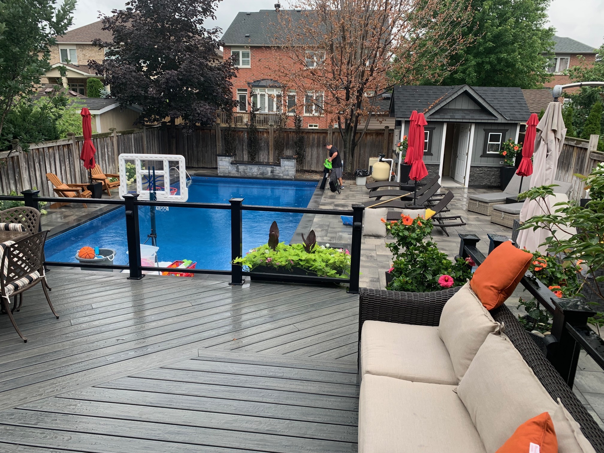 A backyard scene with a pool, patio furniture, vibrant greenery, and a shed. A person tidies up near a house in the background.