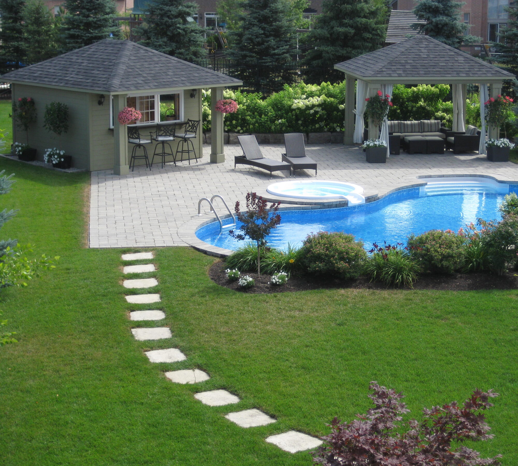 A backyard features a swimming pool, hot tub, two pavilions, lounge chairs, bar area, and manicured lawns with surrounding bushes and trees.
