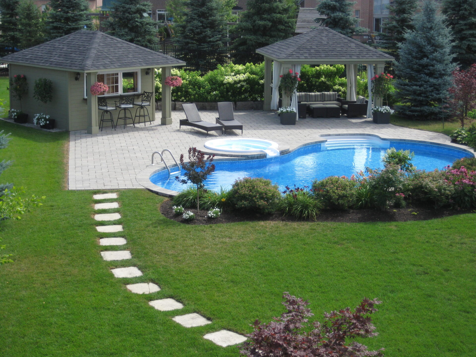 A serene backyard features a swimming pool, hot tub, gazebo, bar area, lush lawn, and vibrant flowers, creating a perfect relaxation spot.