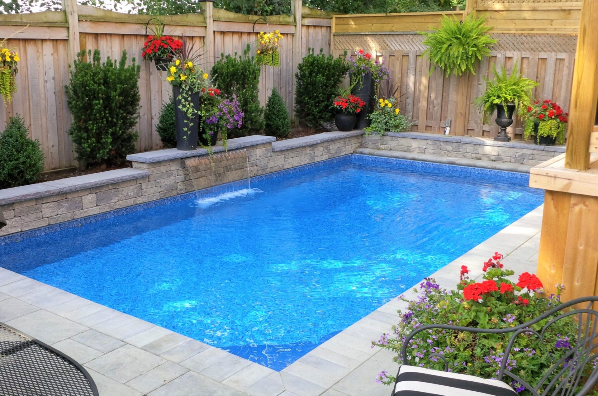 A tranquil backyard features a small blue pool surrounded by stone tiles, vibrant flowers, and a wooden fence, creating a serene atmosphere.