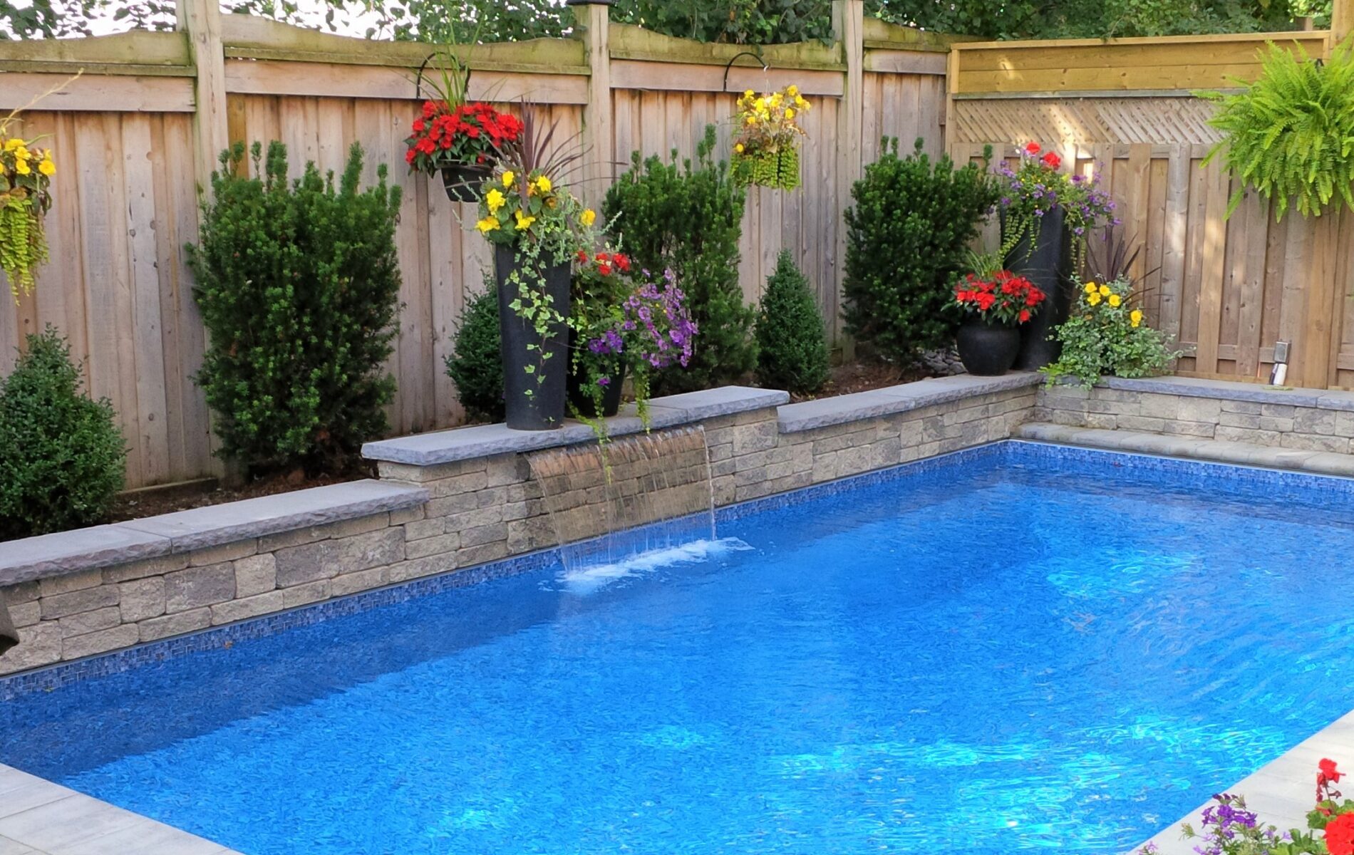 A tranquil backyard features a small blue pool surrounded by stone tiles, vibrant flowers, and a wooden fence, creating a serene atmosphere.