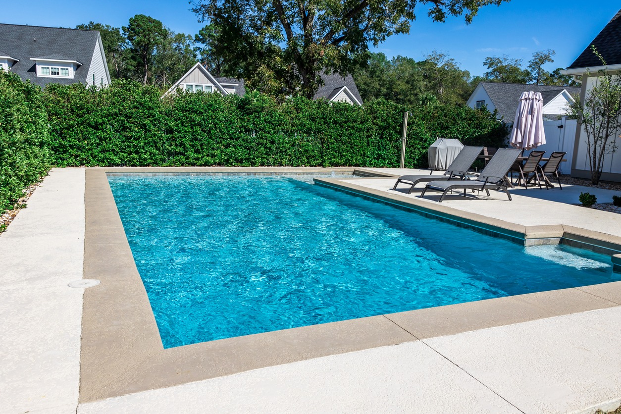 A backyard with a rectangular swimming pool, surrounded by greenery, sun loungers, and patio furniture under blue sky and neighborhood houses.