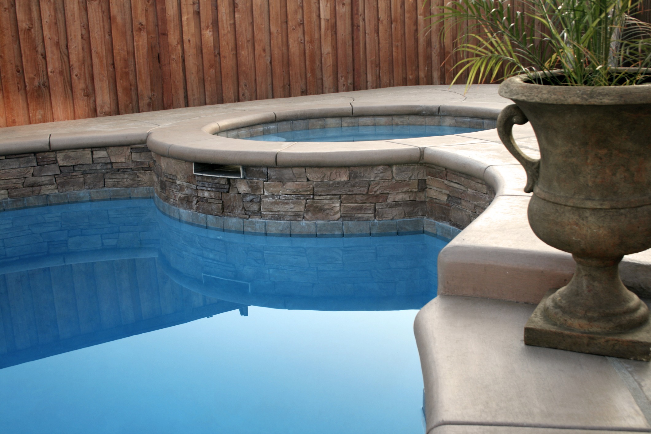 Stone-lined swimming pool with attached hot tub, bordered by wooden fence. Large decorative planter with green plant on poolside.