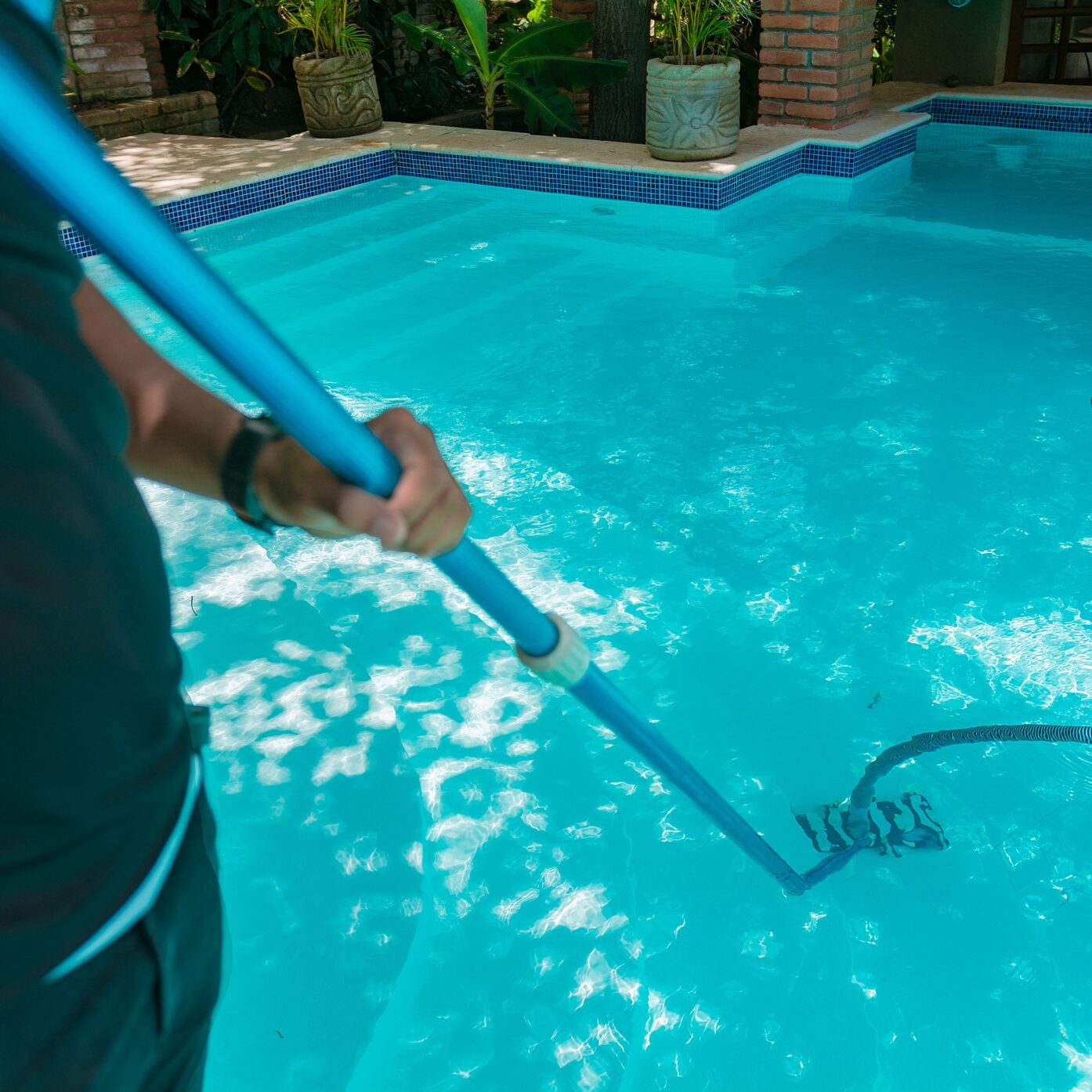 A person is cleaning a swimming pool with a long pole, surrounded by plants and decorative brick pillars.