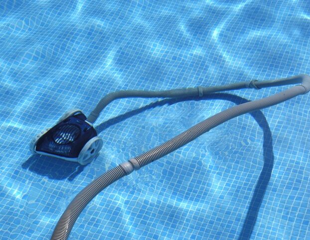A pool vacuum cleaner is submerged, cleaning a blue-tiled swimming pool. Sunlight reflects on the water's surface, creating shimmering patterns.