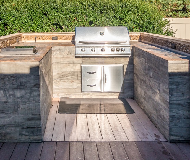 An outdoor kitchen features a modern stainless steel grill, surrounded by stone counters and lush greenery in a residential setting.